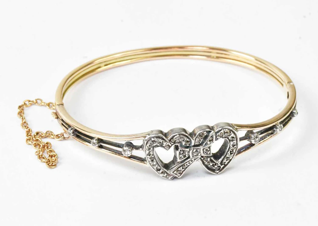 A yellow metal hinged bangle with two white metal hearts entwined with a bow