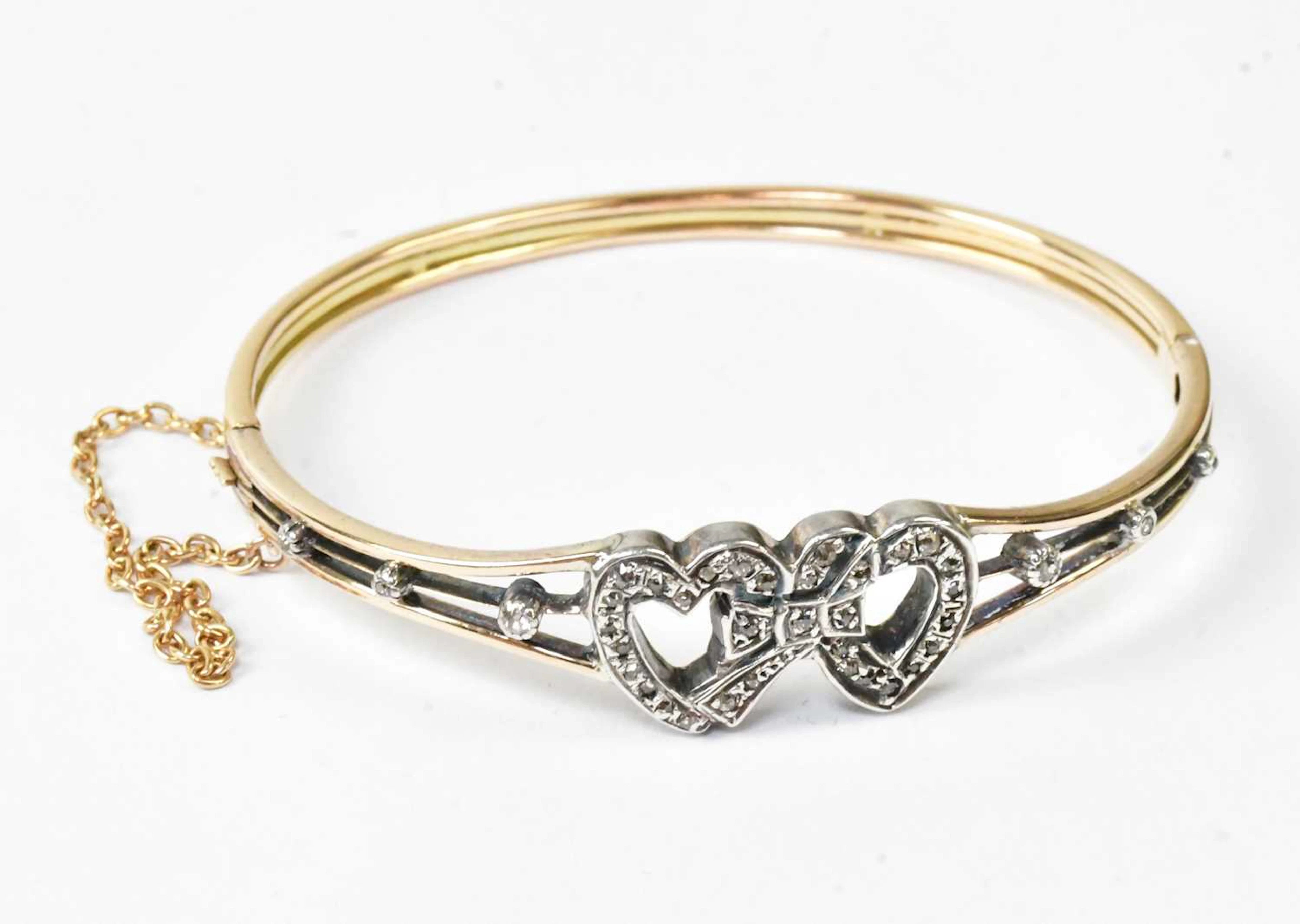 A yellow metal hinged bangle with two white metal hearts entwined with a bow