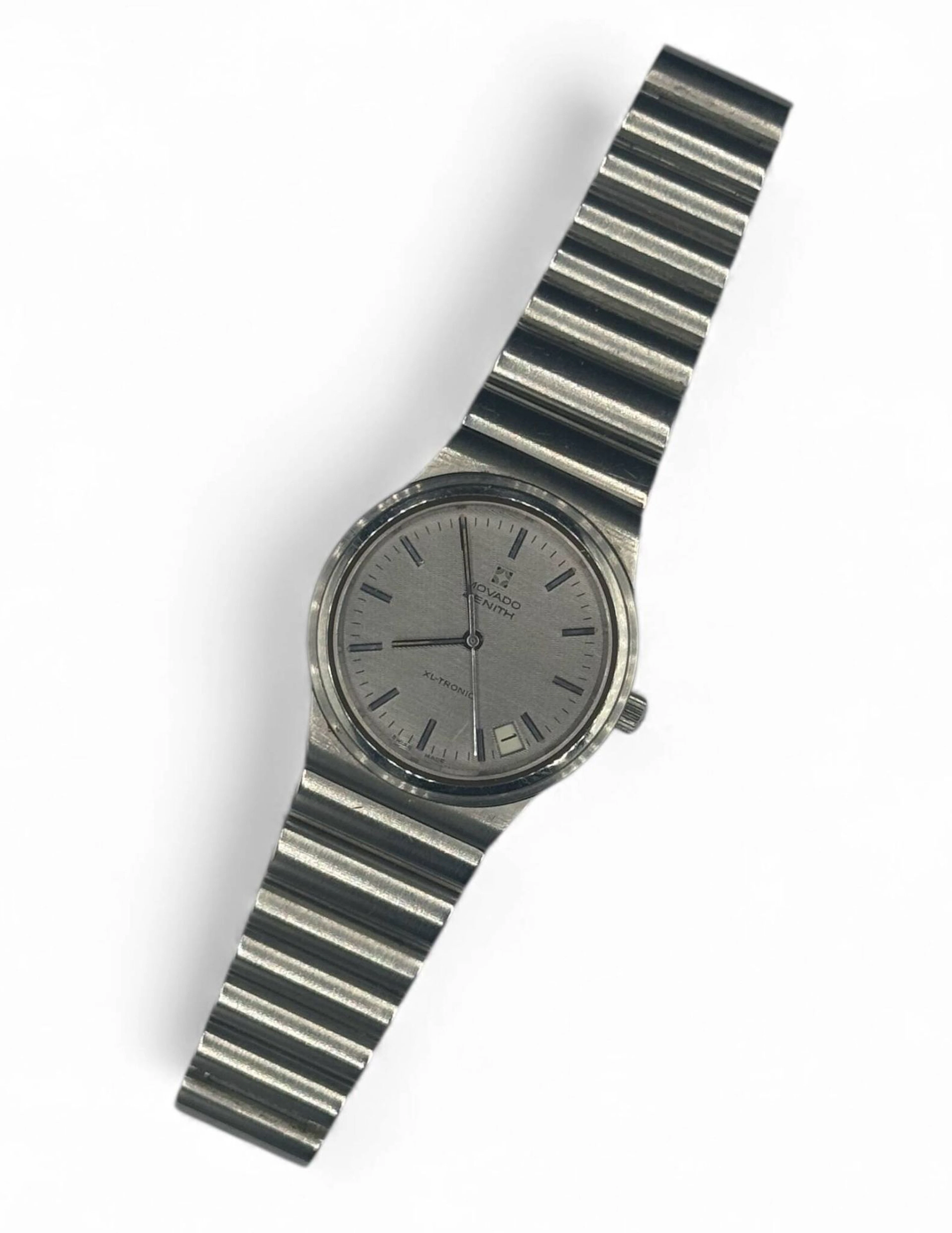 MOVADO ZENITH XL-TRONIC Montre bracelet d’homme en acier, cadran argenté à stries horizontales, index bâtons en appliques argentées et matière luminescente tritium, aiguilles bâtons argentées avec matière luminescente, date entre quatre et cinq heures. Fond en acier vissé, numéroté 01 0050 500, à l’intérieur signé ZENITH mention « MOVEMENT LICENCES BULOVA & PAT ESA », mouvement à diapason signé Zenith base ESA 916 2, numéro 72. Bracelet en acier signé Zenith, boucle déployante.Mouvement non fonctionnel lors de l’expertise, sans garantie de fonctionnement futur et d’état des pièces. Rayures au boitier, au bracelet et au verre. Diamètre 35 mm Vers 1970