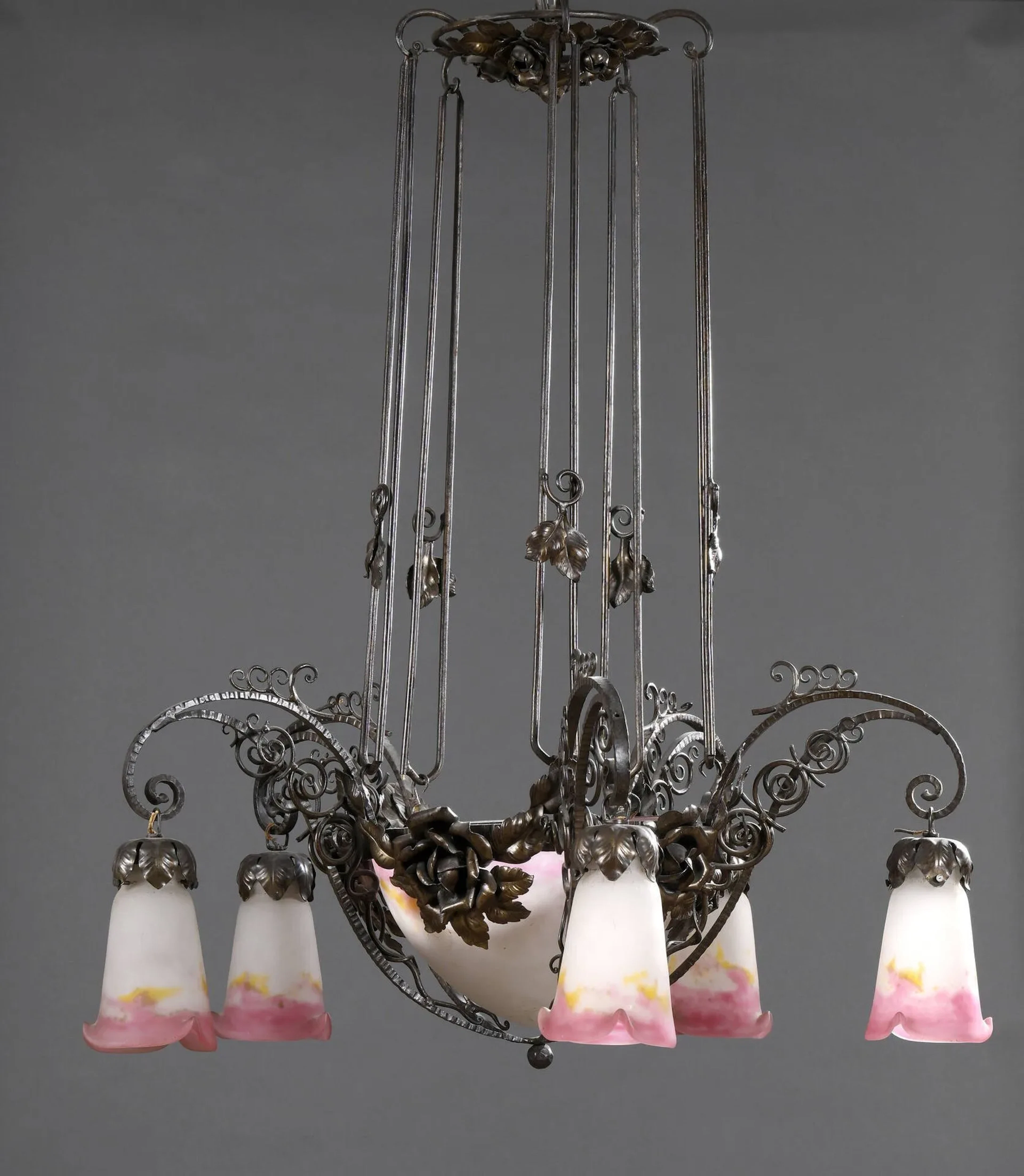 MULLER FRERES – LUNEVILLE Lustre en fer forgé à décor de roses ciselé, martelé et patiné noir à vasque centrale conique et quatre bras de lumières. Cache-ampoule tulipe à bords trilobés en verre blanc tacheté rose et orangé. Signé sur les caches-ampoules. Haut. 90 cm – Diam. 75 cm