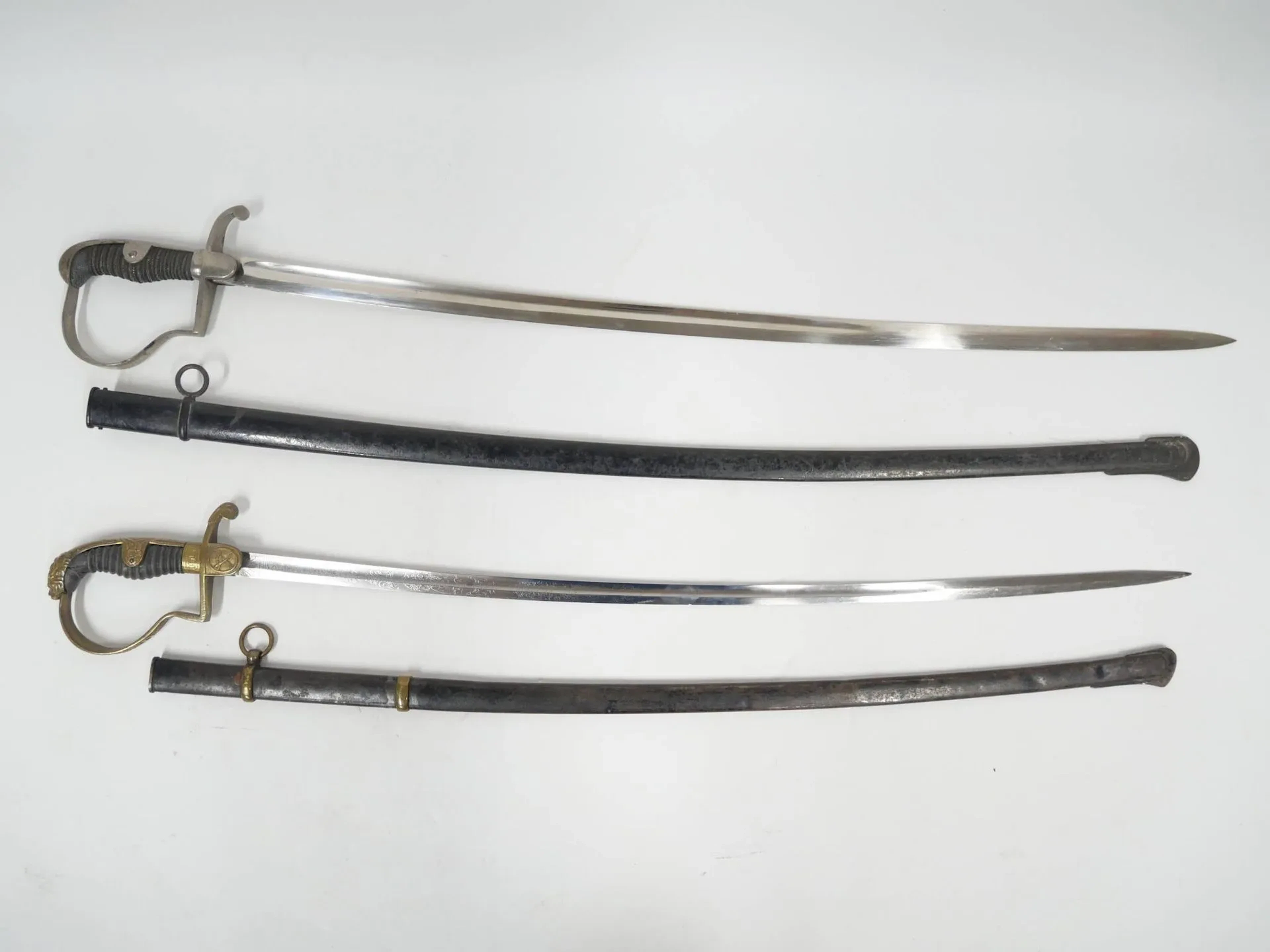 Ensemble de de deux sabres d’officier allemand. Comprenant : -Sabre à la Blücher en laiton ciselé. Calotte à la tete de lion et croisières à deux sabres entrecroisés. Lame courbe à pans creux gravée à l’acide au tiers. Fourreau acier à deux bracelets laiton. - Sabre à la Blücher en fer nickelé. Calotte à la longue queue. Lame courbe à pans creux gravée. Fourreau acier à un bracelet. Allemagne, époque fin du XIXe siècle.