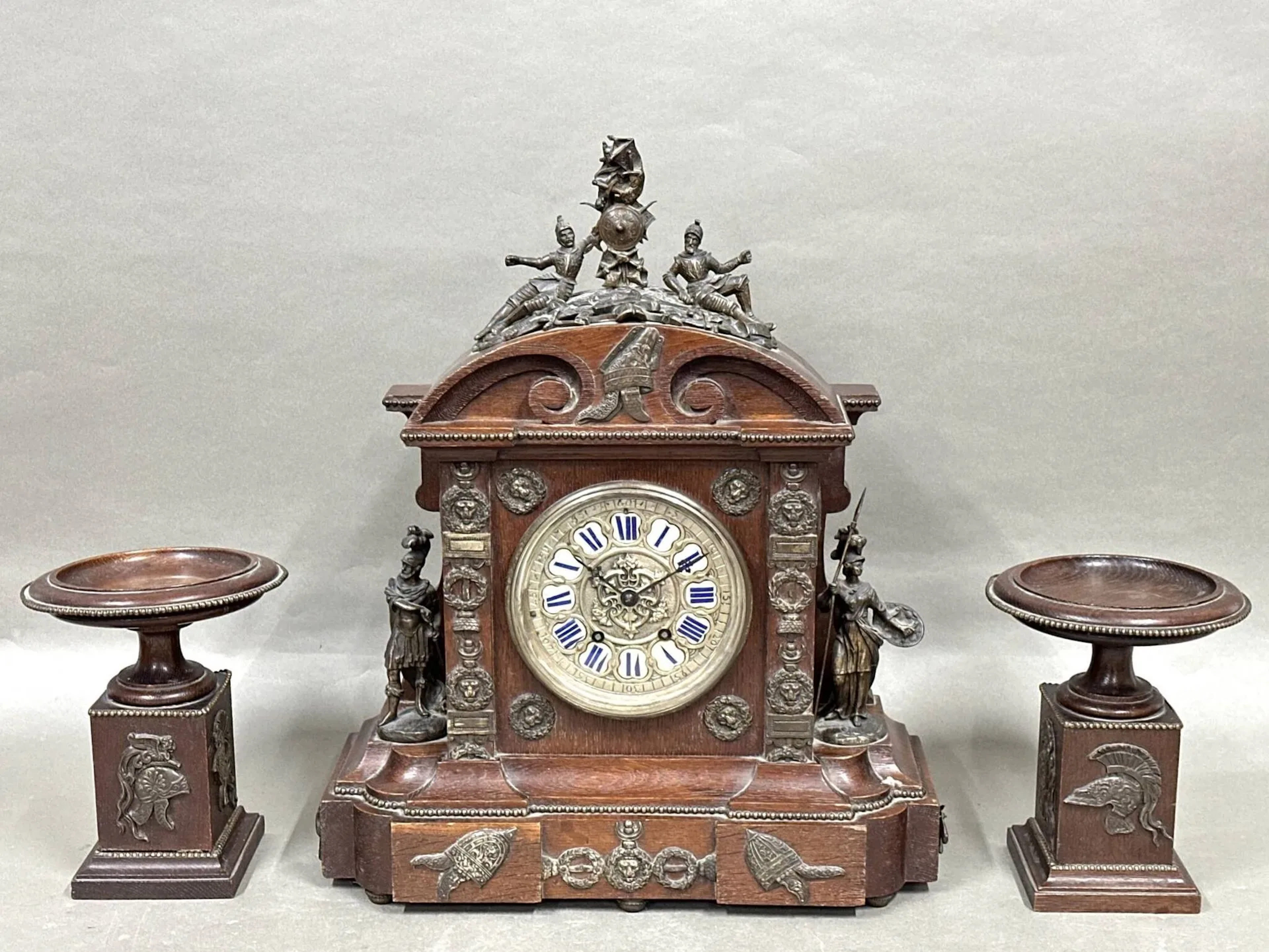 DIELH (dans le goût de) Pendule en bois et placage et ornementation de bronze à décor d'hommes en armure et de frises de laurier. Cadran à cartouches émaillés aux chiffres romains. Accompagnée de ses deux cassolettes. Accidents et manques. Le socle d'une des cassolettes à refixer. Pendule : H. 50 cm - L. 40 cm - P. 16 cm Cassolettes : H. 21 cm.