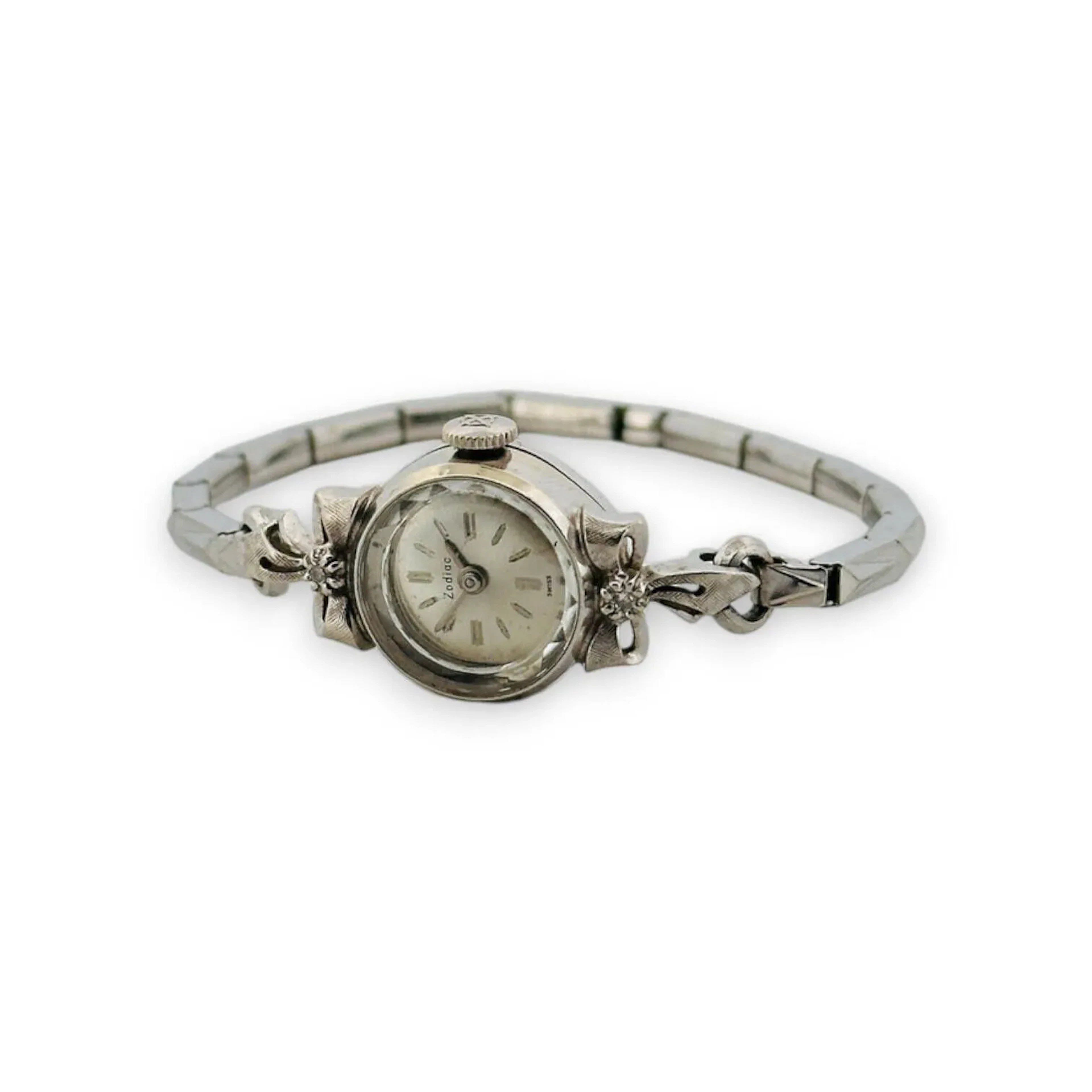 ZODIAC Montre bracelet de dame, boitier rond en or 14K (585), lunettes à décor rubané piqué d'un petit diamant, cadran argenté, index bâtons formant les heures, mouvement mécanique, bracelet intégré extensible en métal argenté. Diam. : 15 mm - Poids brut : 12.19 g.