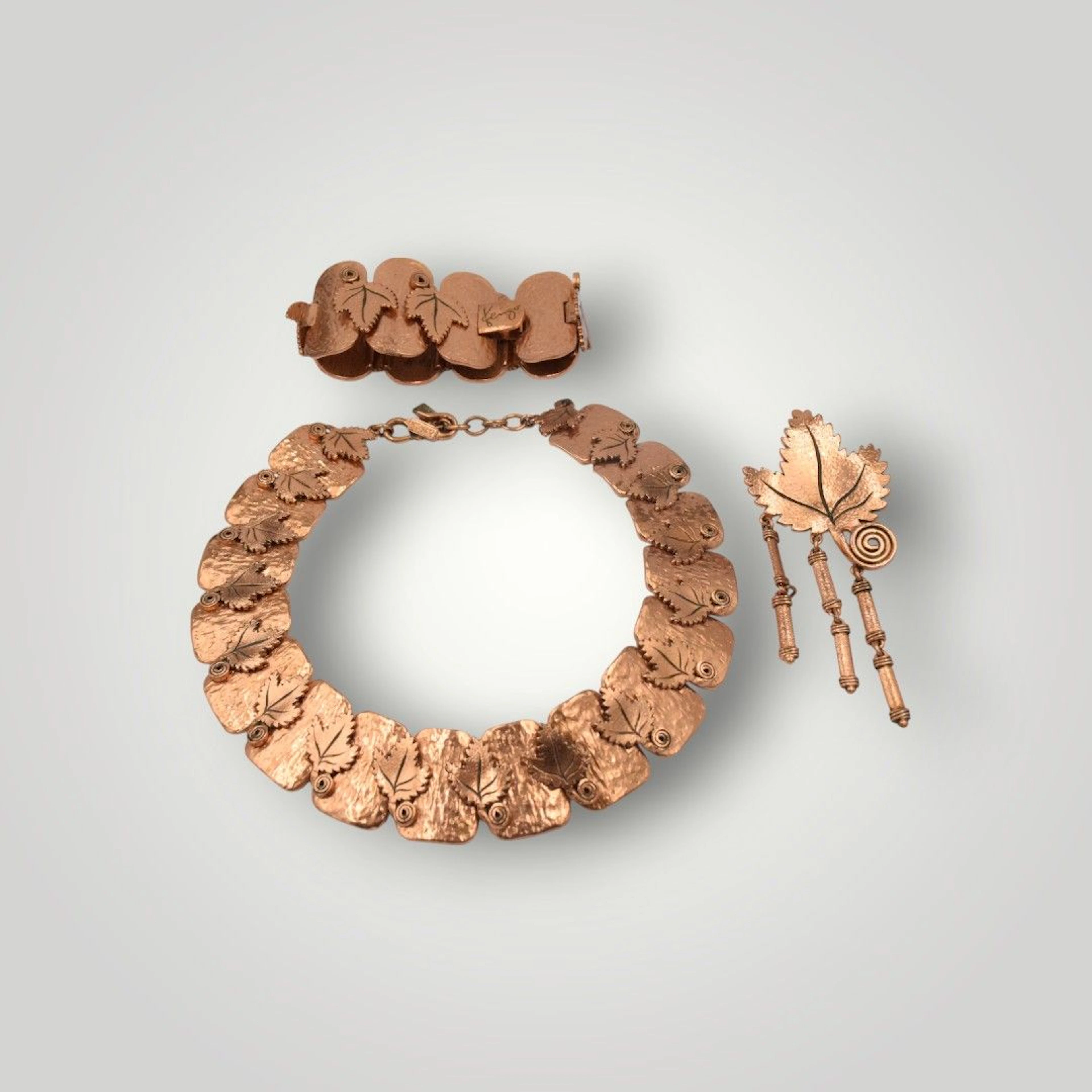 KENZO Paris Parure en métal cuivré à décor de feuilles de vignes et plaques martelées comprenant : - Une broche formant une feuille de vigne supportant trois pampilles. - Un bracelet fermoir clip reprenant les mêmes motifs. - Un collier en métal cuivré fermoir en S reprenant les mêmes motifs.