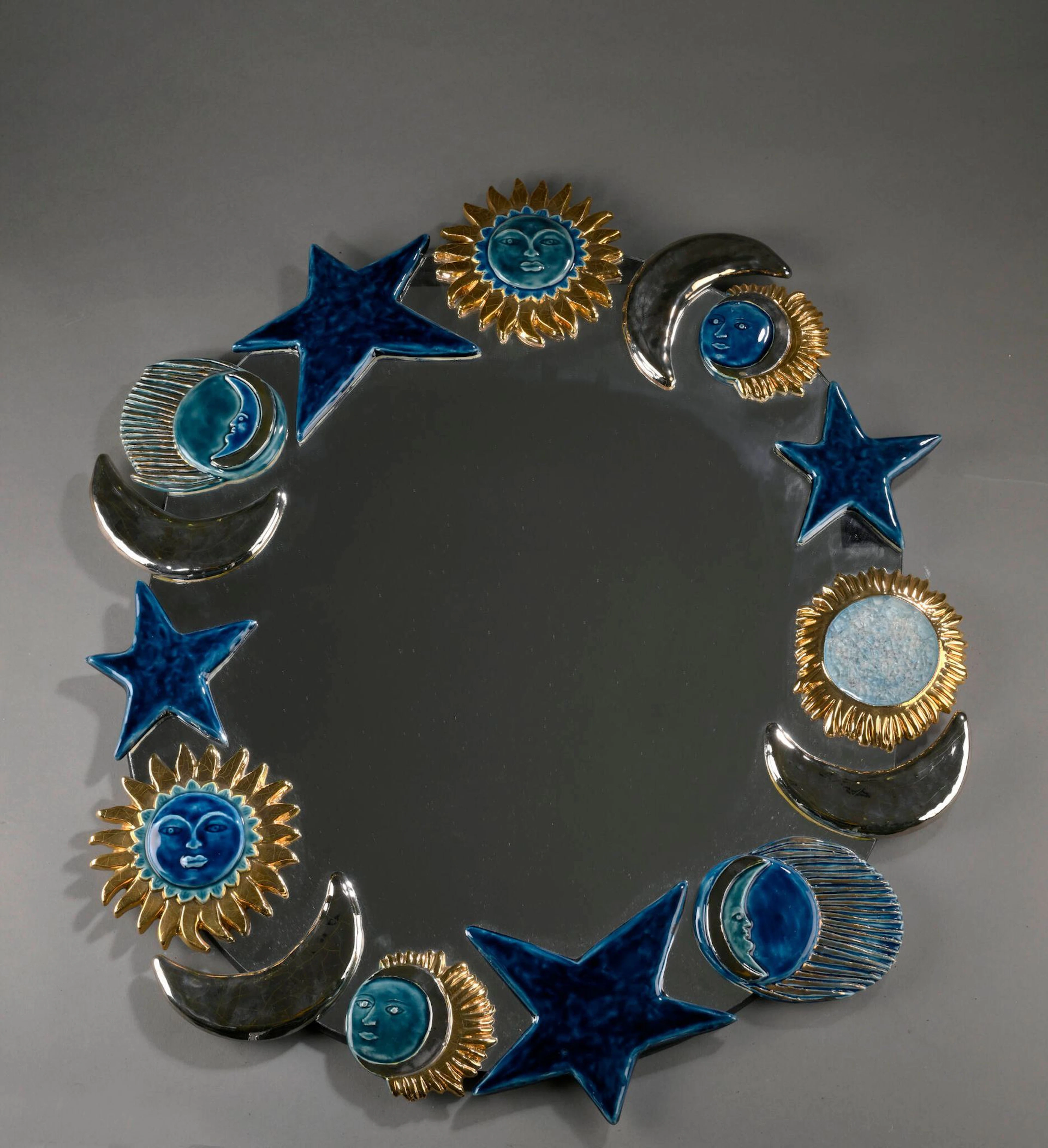 Marion de CRECY (1954) Miroir mural de forme ovale à décor de soleils, lunes et étoiles Épreuve en céramique émaillée or, argent, bleu et verre cristallisé Non signé H. : 73 cm. – L. : 66 cm.