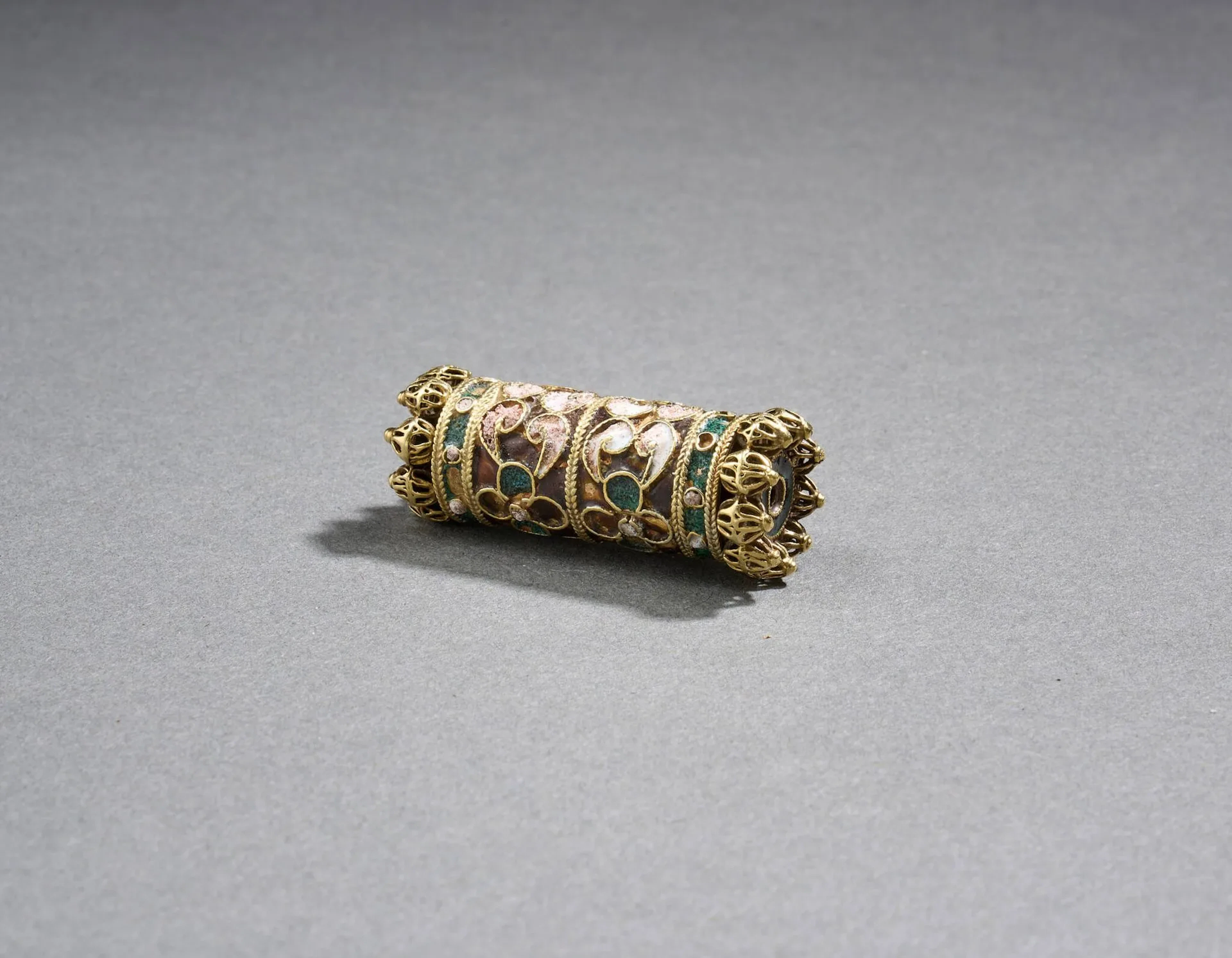 Élément qannuta de collier dit al-haiathe, Nord du Maroc, probablement Tétouan, XVIIIe siècle Élément tubulaire en or à décor d’émaux polychromes cloisonnés dessinant des motifs végétaux à quatre feuilles lobées, une frise de petites sphères filigranées et ajourées sur chacun des côtés. S’ouvre aux deux extrémités, formant boîtier. H : 4,5 cm ; PB. 16 g Petits accidents à l’émail. Cet élément cylindrique, appelé qannuta de l’arabe qannût(a) / « portion de roseau prise entre deux nœuds » sans doute par analogie formelle, complétait une parure de cou aujourd’hui disparue. Le type de collier qu’il rythmait, généralement en alternance avec des boules d’or filigrané ou des chapelets de petites perles baroques de part et d’autre d’un pendentif central, reprend un prototype connu dans la joaillerie nasride des XIVe – XVe siècle comme en attestent le collier du trésor de Mondujar au Musée archéologique national de Madrid (51033) ou la parure démontée du Metropolitan Museum de New York (17.190.161a-j). L’immigration d’orfèvres juifs de l’Espagne vers l’Afrique du Nord, essentiellement au moment de la Reconquista, a permis ce transfert de modèles et de techniques, dont l’émail cloisonné et le filigrane. La forme tubulaire de ces perles se démontant comme un petit boîtier montre également une continuité culturelle, s’inscrivant dans la tradition des boîtiers talismaniques communs aux cultures juive, musulmane et mêmes à quelques civilisations antiques, qui jouaient un rôle protecteur grâce aux formules religieuses qu’ils renfermaient. Dans les parures nasrides puis marocaines, ce rôle prophylactique est abandonné, laissant place à une fonction purement ornementale. Peu d’exemples de qannuta émaillées nasrides ont survécu. Une pièce de la Hispanic Society of America à New York (R3402) est attribuée à un travail espagnol du premier quart du XVIe siècle et les qannuta du collier du MET précité sont attribuées à la fin du XVe siècle ou au début du XVIe siècle. Les pièces marocaines plus tardives comme celle-ci, qui montrent une vraie assimilation des techniques ancestrales, sont plus fréquentes. On pourrait citer une qannuta de l’Institut du Monde Arabe attribuée à Tétouan au XVIIIe siècle comme deux autres sur un collier encore complet du Victoria and Albert Museum de Londres (607:1, 2-1902). Bibliographie GONZALEZ, Valérie, Emaux d’al-Andalus et du Maghreb, Edisud, Aix-en-Provence : 1994, pp. 128, 129, 180, 189, 190. RABATE Marie-Rose, GOLDENBERG, André, Bijoux du Maroc, Edisud, Aix-en-Provence : 1999, p.212.