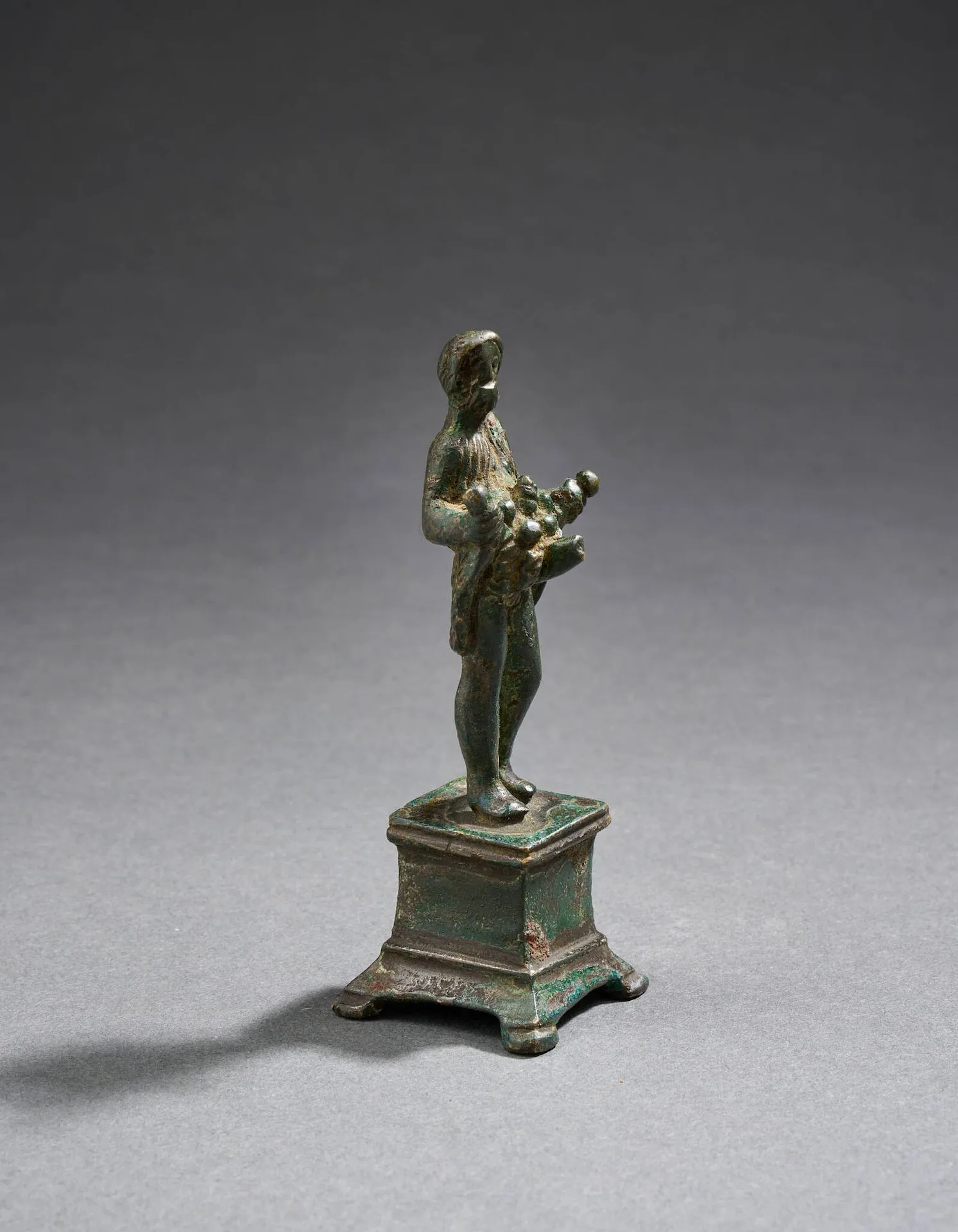 Statuette de Priape vêtu d’une tunique courte qu’il relève, dévoilant son sexe en érection. Dans le pli du vêtement se trouvent des fruits. Il repose sur une base quadripode. Bronze à patine vert bleuté. Art romain, IIe/IIIe siècle. Hauteur : 8,2 cm