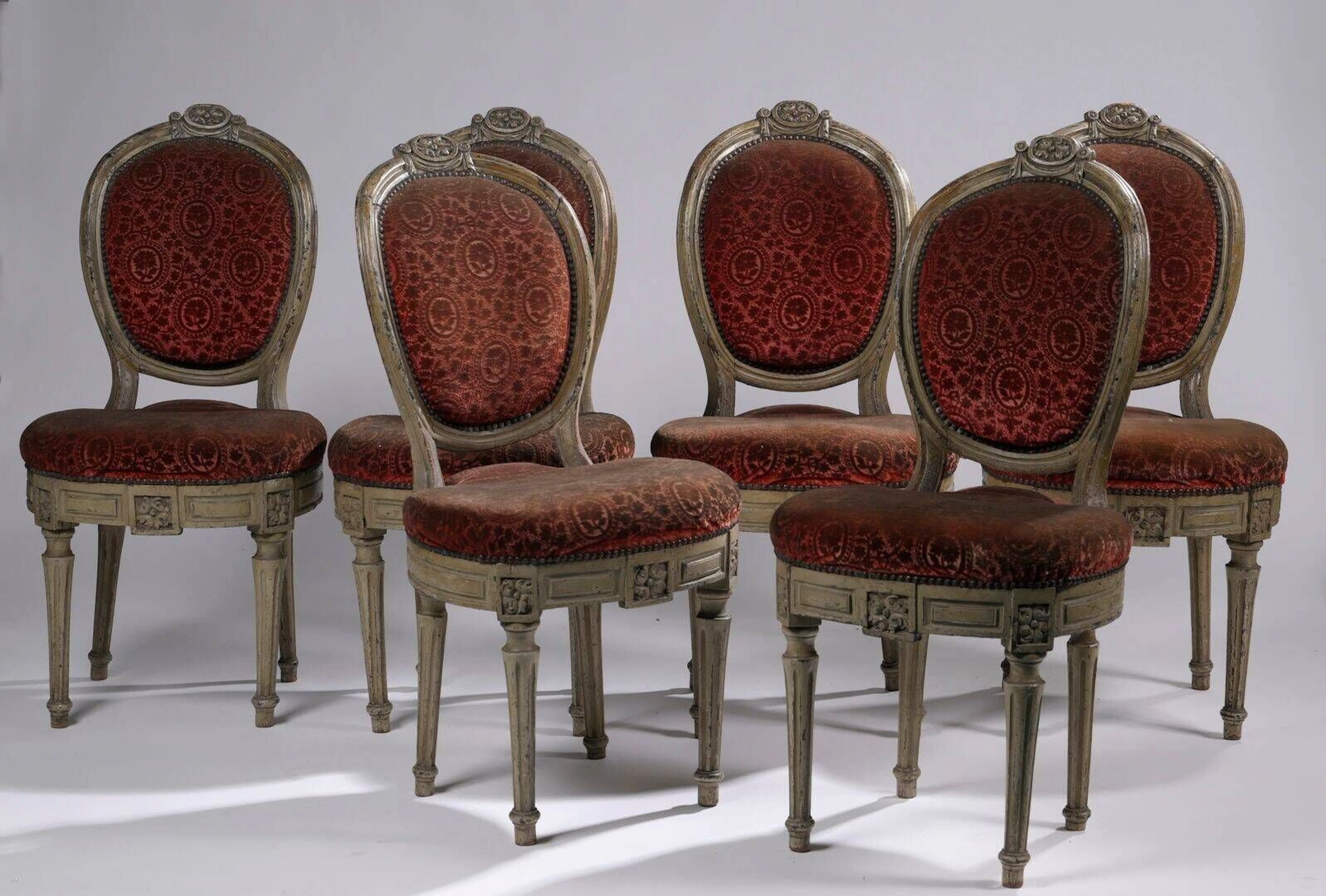 Suite de six chaises cabriolet anciennement cannées à dossier médaillon en bois relaqué gris mouluré et sculpté de quartefeuilles dans des médaillons, celui du dossier entouré de draperies, l’assise circulaire, les pieds fuselés cannelés. Epoque Louis XVI (accidents, renforts, restaurations et parties refaites) Garnitures de velours frappé coq de roche