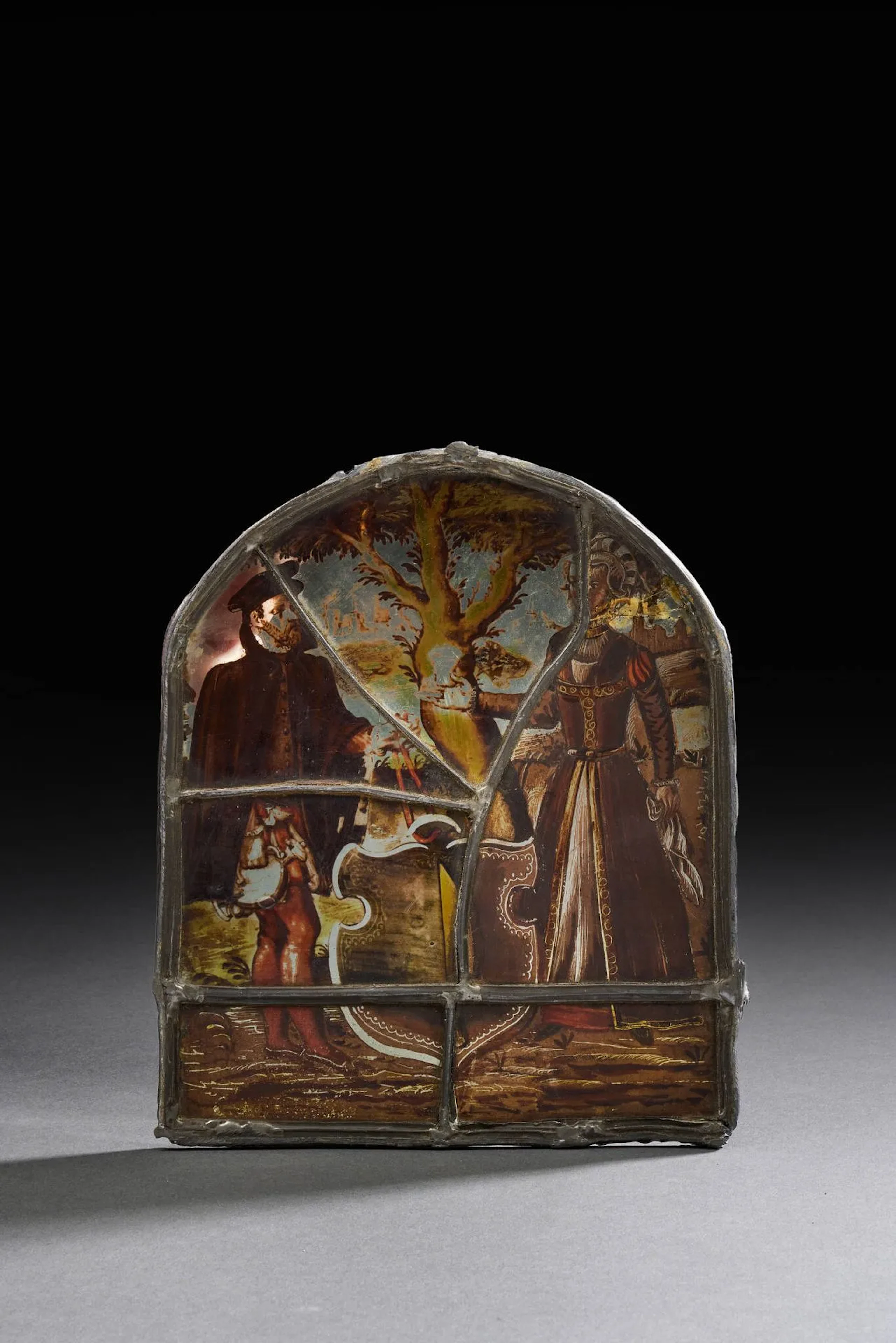 Vitrail de forme cintrée peint en grisaille de brun rehaussée de jaune d’argent et d’orange représentant un couple en pied : la femme tend un verre à son époux qui tient une gourde dans sa main droite et les liens d’un écu meublé d’un couteau de l’autre ; cet écu au contour échancré est posé devant un arbre avec paysage au second plan Pays alémanique, en partie du XVIe siècle Hauteur : 21,5 cm – Largeur : 18 cm (personnage féminin refait anciennement, petite cassure et plombs de casse)