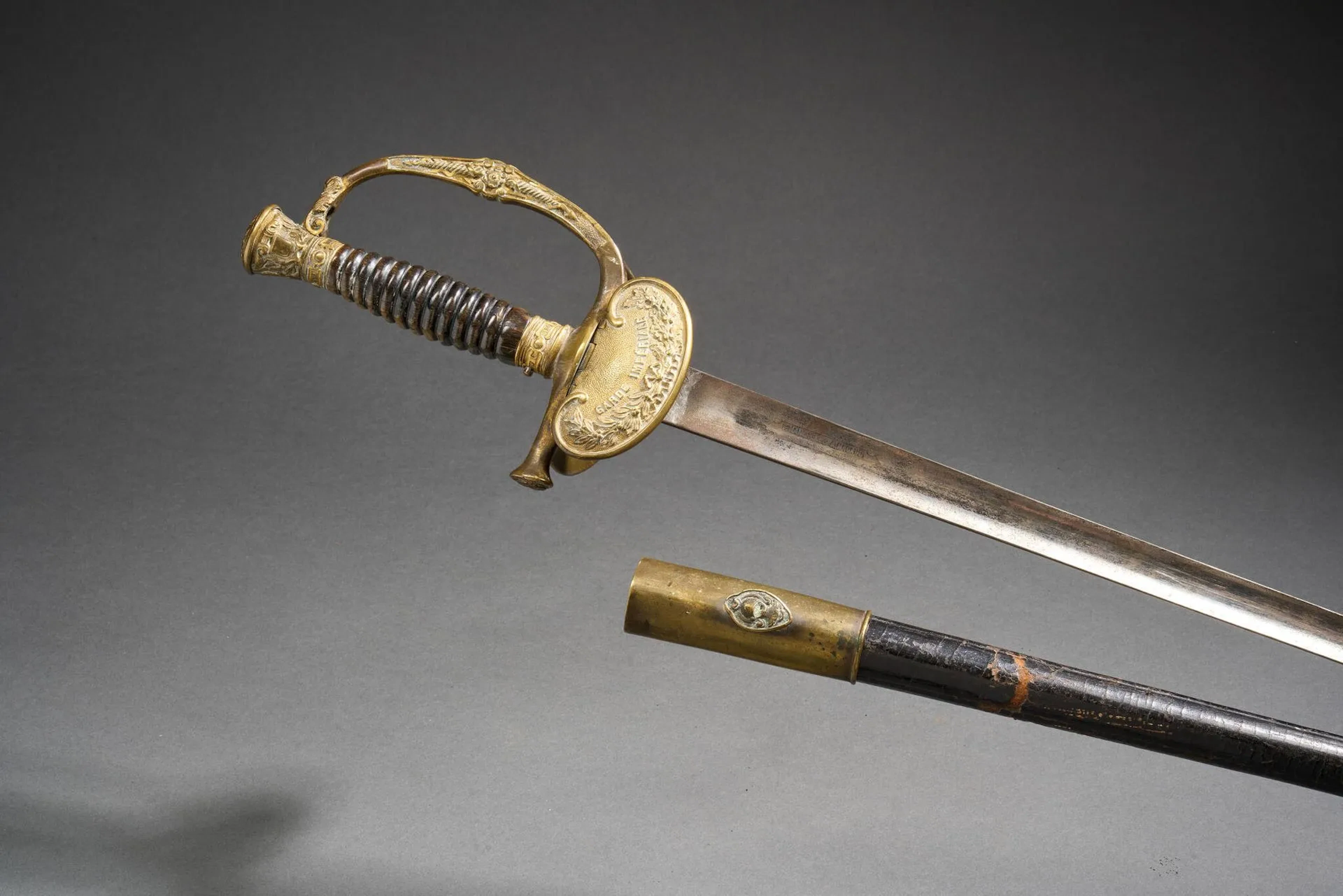 Épée d’officier de la Garde Impériale modèle 1860. Monture en laiton ciselé. Pommeau à l’aigle, arc de jointure au bâton de foudre avec le chiffre « N » au centre et quillon droit à la feuille d’acanthe. Clavier aux grandes armes impériales et contre-clavier à pompe. Fusée en corne noircie (manque le filigrane). Lame droite de section triangulaire marquée « F.Manchon & A.Mangaud Rue Richelieu 12 Paris. Fourreau en cuir conforme au règlement (manques et accidents au cuir). Longueur totale : 96 cm. France, Époque Second Empire.