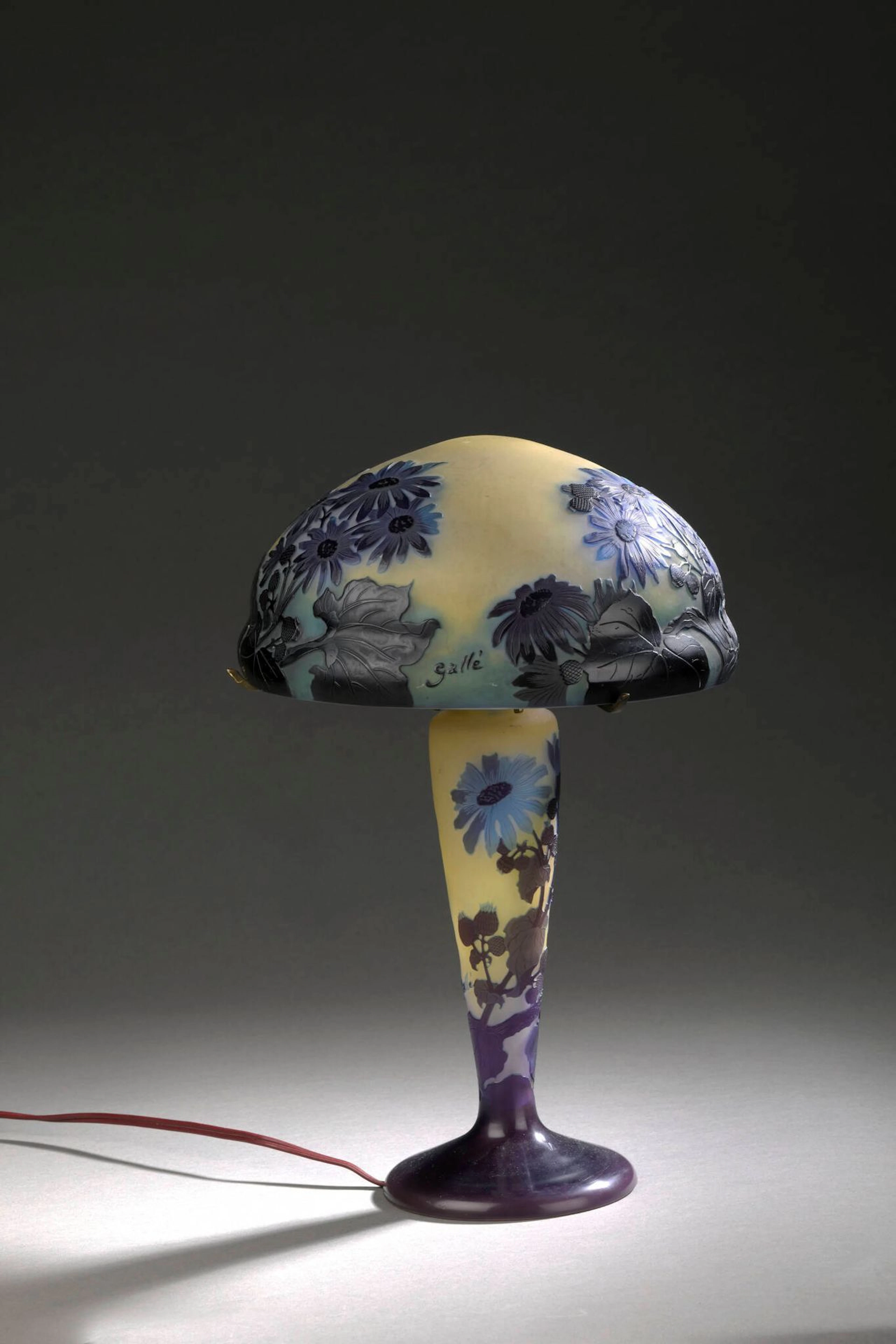 ÉTABLISSEMENTS GALLÉ (1904-1936) Lampe de table à pied conique épaulé sur base circulaire et abat-jour champignon. Épreuves en verre violet et bleu sur fond jaune. Décor de gerbera gravé en camée à l’acide. Système à trois bras en laiton (petites égrenures au col). Signé sur le pied et l’abat-jour. Haut. 36 cm (totale) Bibliographie : Alastair Duncan & Georges de Bartha, Gallé Lamps, Antique Collectors’ Club, Woodbridge, 2014, modèle similaire référencé et reproduit p. 117, pl. 7.7.