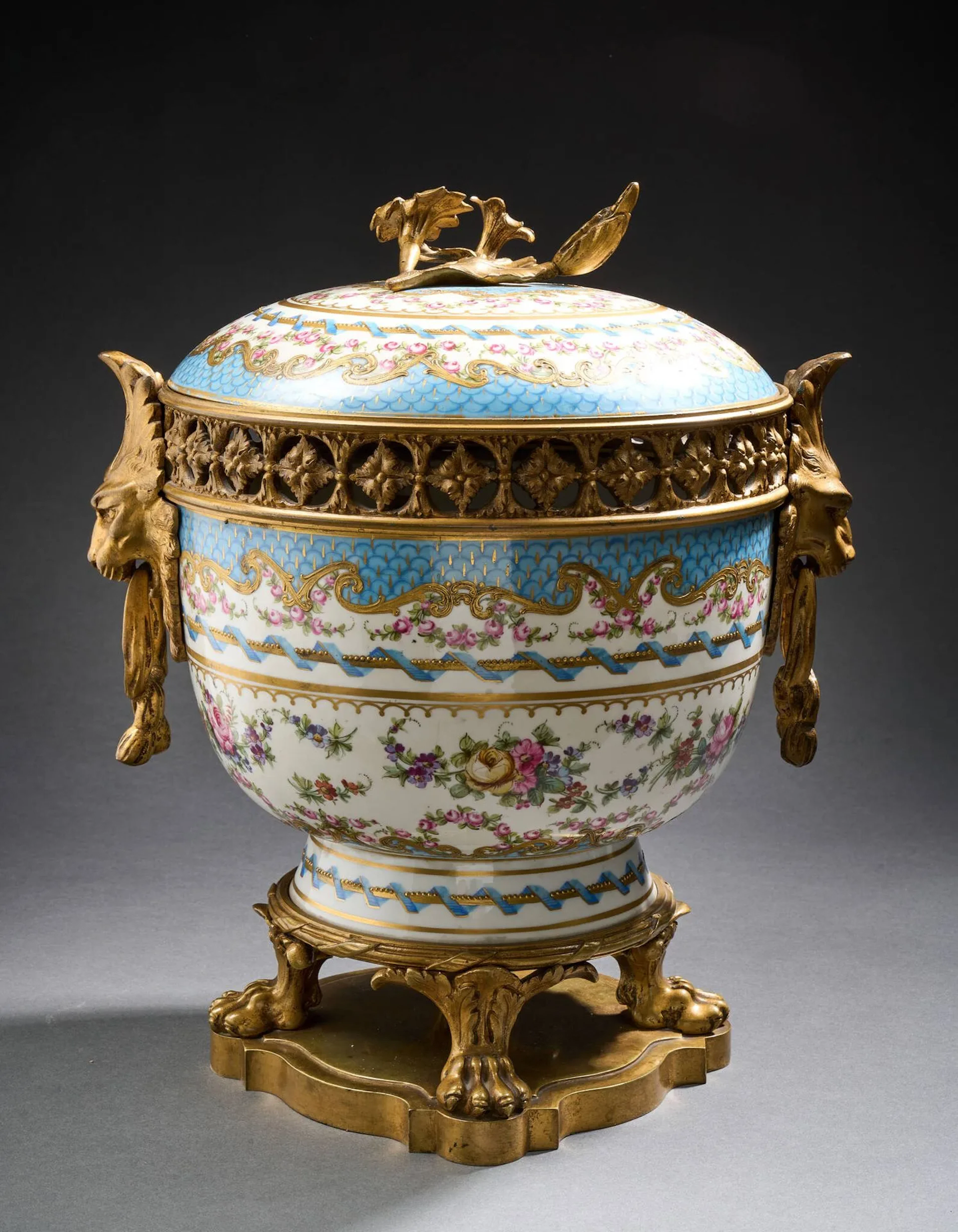 SÈVRES, dans le goût de. Important pot-pourri circulaire et son couvercle en porcelaine à décor polychrome et or alterné de frises d’écailles, vagues, roses et filets rubanés bleus. Monté en bronze doré, reposant sur un socle à quatre pieds en forme de pattes de lion, les anses appliquées d’un décor de mascaron, la prise en trompe-l'œil de fleur. Seconde moitié du XIXe siècle. Portant de fausses marques de la manufacture de Sèvres au revers. H. 35 x L. 24,5 cm.