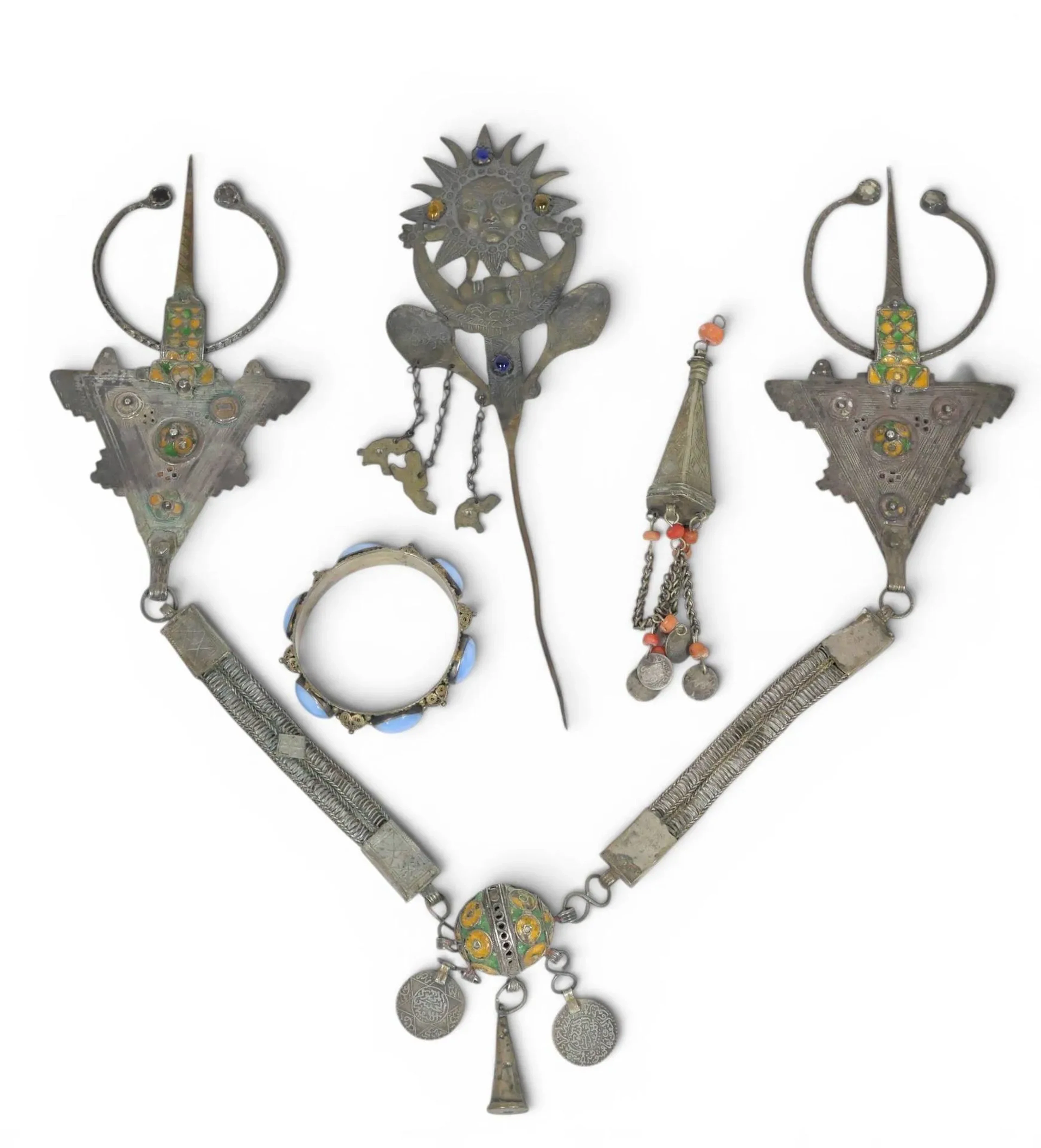 Parure pectorale, Maroc, Anti-Atlas occidental, Région de Tiznit, début XXe siècle Parure en argent composée d’une paire de fibules tizerzaï à contours crénelés, décor ciselé, ajouré et de cabochons émaillés jaune et vert, leur anneau à cabochons de verre transparent ou coloré, reliées par une large chaîne à une boule centrale émaillée dite tagmout soulignée d’une pendeloque conique et deux dirhams datés 1310 H / 1892 et 1321 H /1903. Longueur : 69 cm Poids brut : 300 grammes Oxydation, petites déformations, petits sauts d’émail. On joint un bracelet à cabochons bleus, une pendeloque en argent à pampilles de monnaies et perles de corail (poids brut : 35 grammes) et une épingle en métal découpé à décor de soleil et de croissant de lune.