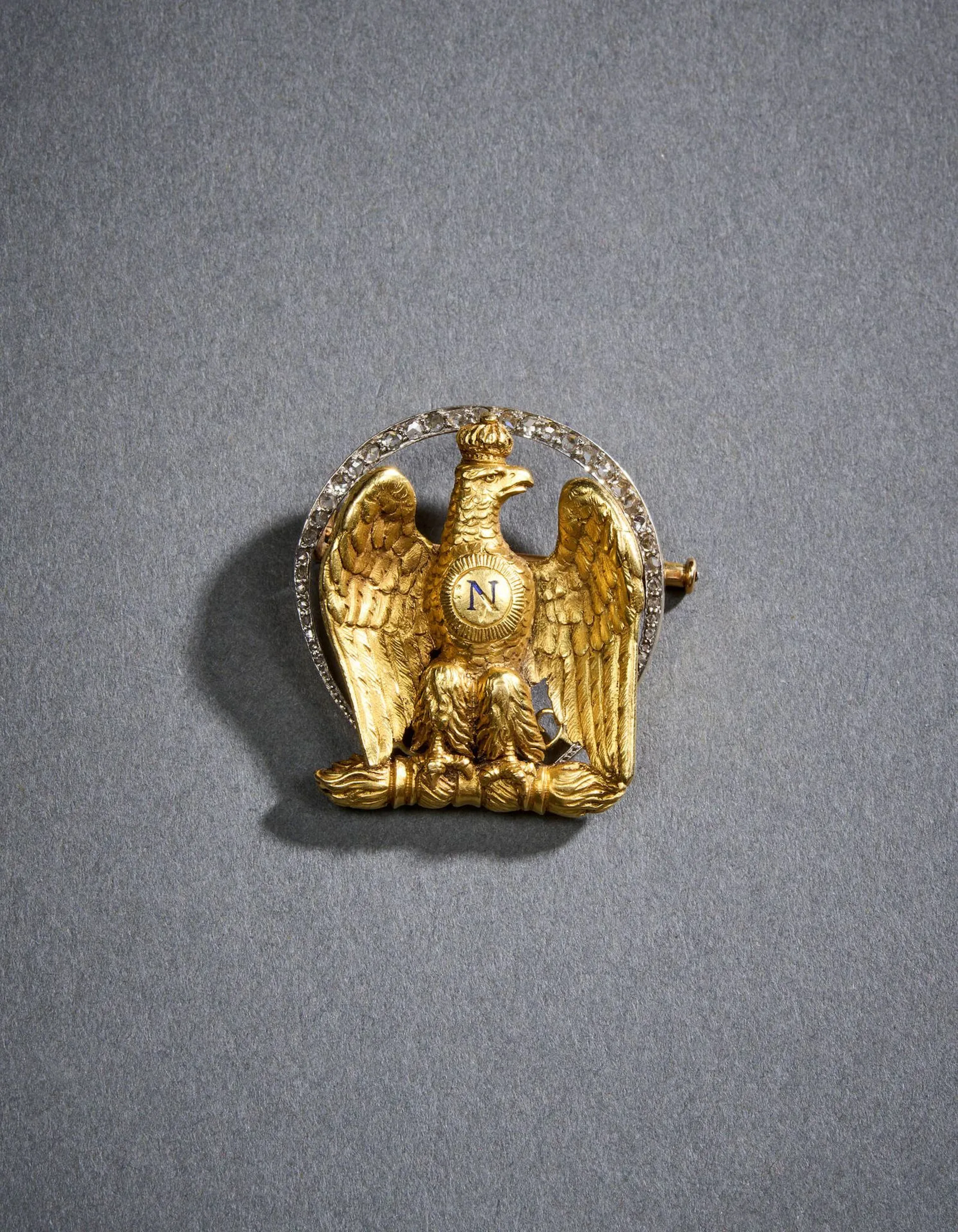 Broche bonapartiste en or (750 millièmes) ciselé figurant une aigle couronnée dressée sur le foudre impérial et centrée du N de Napoléon émaillé bleu, entourée d’une chute de diamants taille rose et sertis sur platine. Fixation par épingle basculante. Petit manque d’émail. Fin du XIXe siècle. Sans poinçon apparent. H. 3,6 x L. 3,4 cm. Poids brut : 28,5 g.