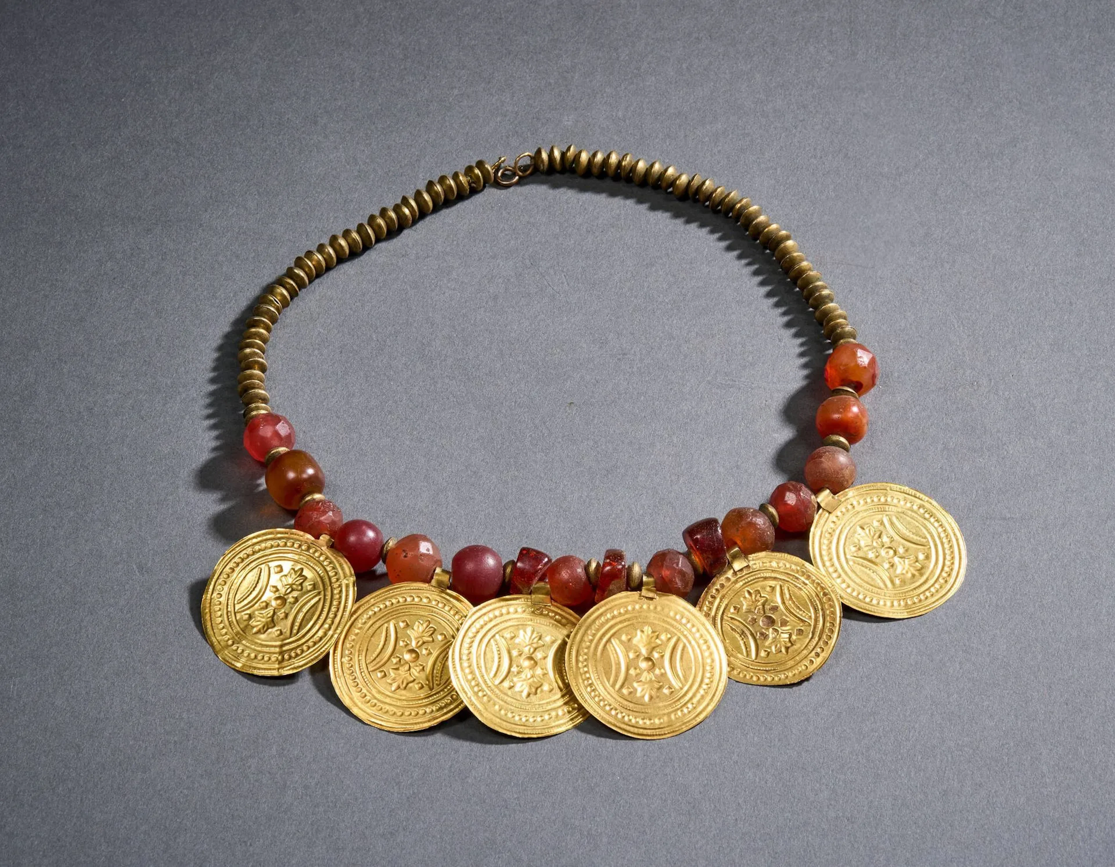 Collier draperie en or (750 millièmes), argent et cornaline, à décor alterné d’une suite de 6 disques en or repoussé à décor de frises et motifs géométriques stylisées, de 16 perles en cornaline, certaines facettées, et de perles légèrement aplaties en vermeil (800 millièmes), le fermoir en or (385 millièmes). Petits chocs. fermoir métal. Travail égyptien du XIXe siècle, dans le goût antique. Sans poinçon apparent. L. 45 cm. Poids brut : 64,8 g.
