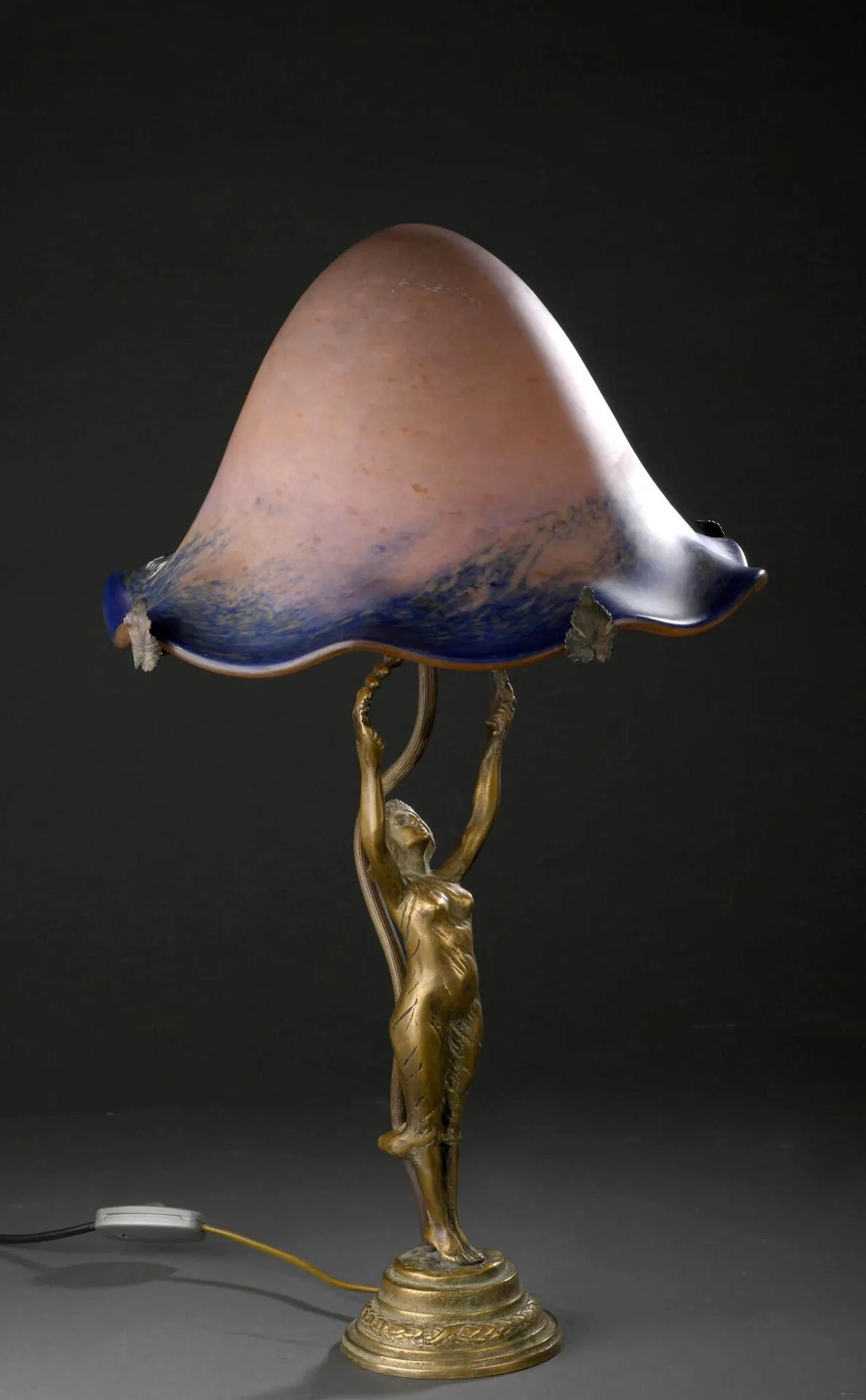 Charles SCHNEIDER (1881 – 1953) – LE VERRE FRANÇAIS & LES LUMIERES DE NANCY (bronzier) « Jeune fille aux bras levés à la guirlande ». Lampe de table en bronze doré patiné à quatre tiges de maintien tubulaire retenant un abat-jour conique à bords chantournés en verre rosé marmoréen bleu et vert (montage postérieur). Signé sur la base et sur l’abat-jour. Haut. 58 cm (totale)