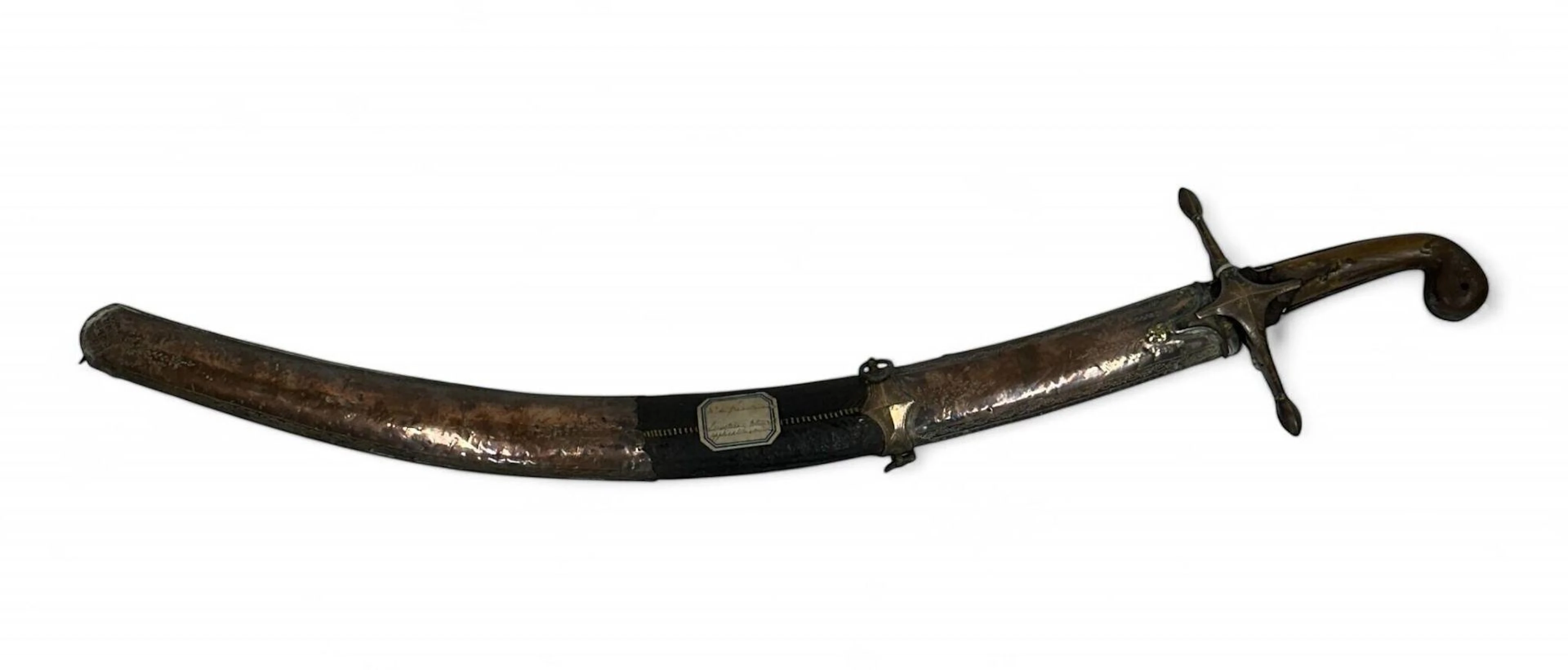 Sabre oriental dit Pala. Monture en forme de crosse, en corne blonde et alliage cuivreux. Croisière cruciforme à deux longs quillons droits, ciselée à décors géométriques et floraux. Plaquettes en corne blonde rivetées et percées pour passage de la dragonne. Pièce de garde désolidarisée, possiblement rapportée bien que parfaitement ajustée. Lame type Pala à dos plat puis fort contre-tranchant. Elle est damasquinée à décors de fleurs et caractères en arabe dans un cartouche. Fourreau à âme en bois recouvert d’une basane, deux garnitures en cuivre découpées et décorées aux points. Bracelet sous la chape, à deux anneaux de suspente. Présence d’une ancienne étiquette inscrite « Mr de Gréaulme / Cimeterre applications cuivre ». Longueur totale : 84 cm. Flèche : 7 cm. Empire ottoman, époque fin du XVIIIe, début du XIXe siècle.