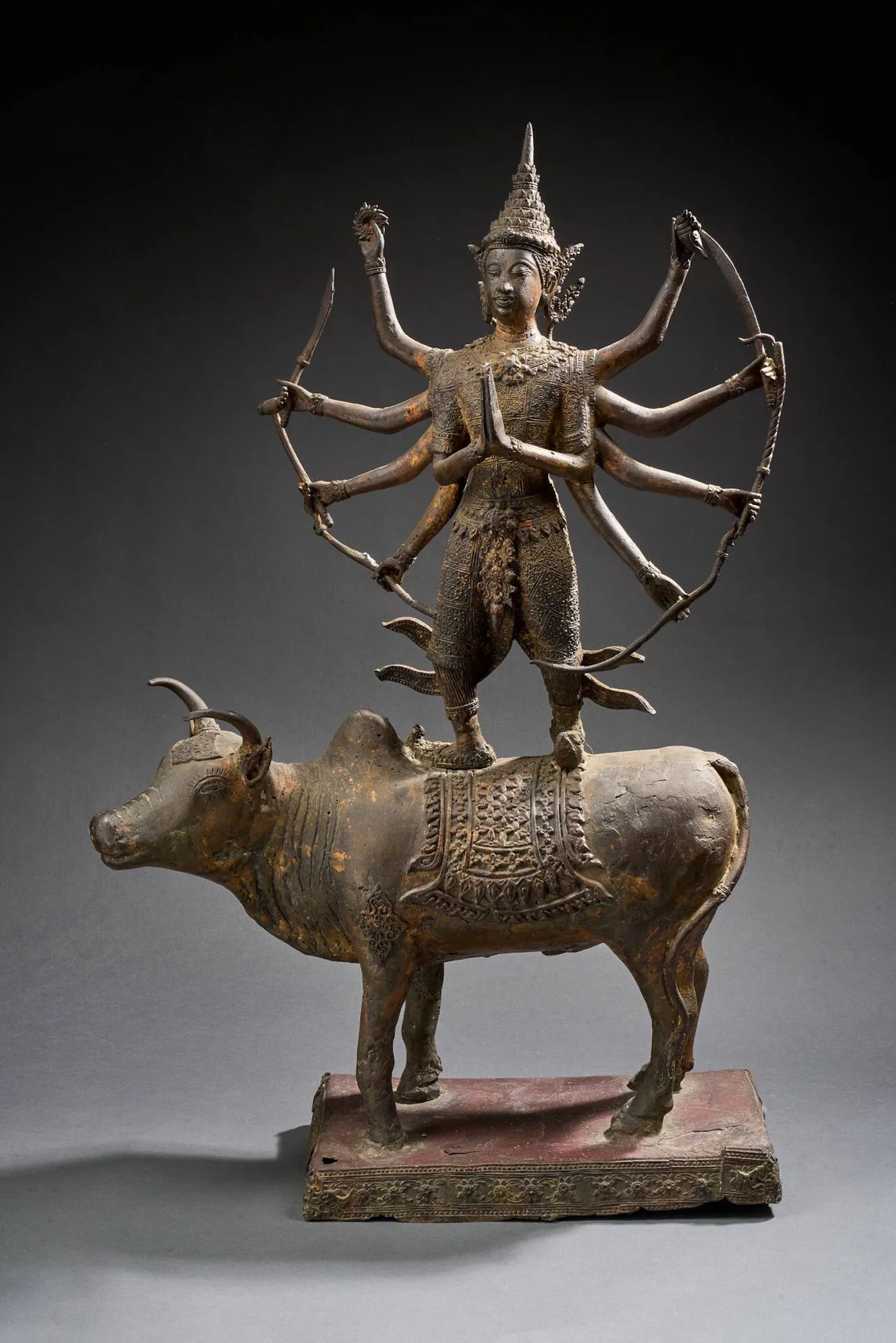 THAILANDE, Ratanakosin - Vers 1900 Grande statue de divinité à dix bras debout sur un buffle posé sur un socle en bronze laqué or et rouge. Tenant dans ses mains des attributs (conque, épée, arc, perle massue), les mains originales en anjali mudra. (Accidents et restaurations). H. 79 cm