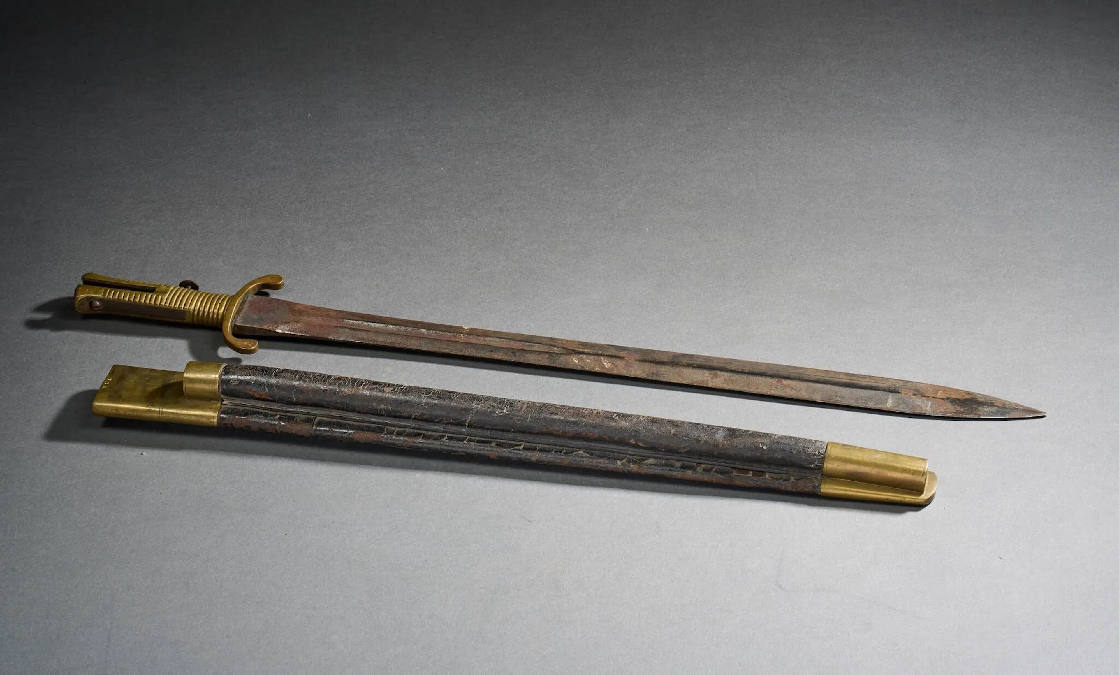 Russie impériale Glaive baïonnette modèle 1843. Fabrication Malherbe à Liège. Monture en laiton avec bouton de verrouillage en fer, double quillon courbe. Lame droite à gorge puis arête médiane marquée au talon PJ. Malherbe à Liège. Fourreau en cuir à deux garnitures laiton. Russie, époque milieu du XIXe siècle Longueur totale : 66,5 cm.