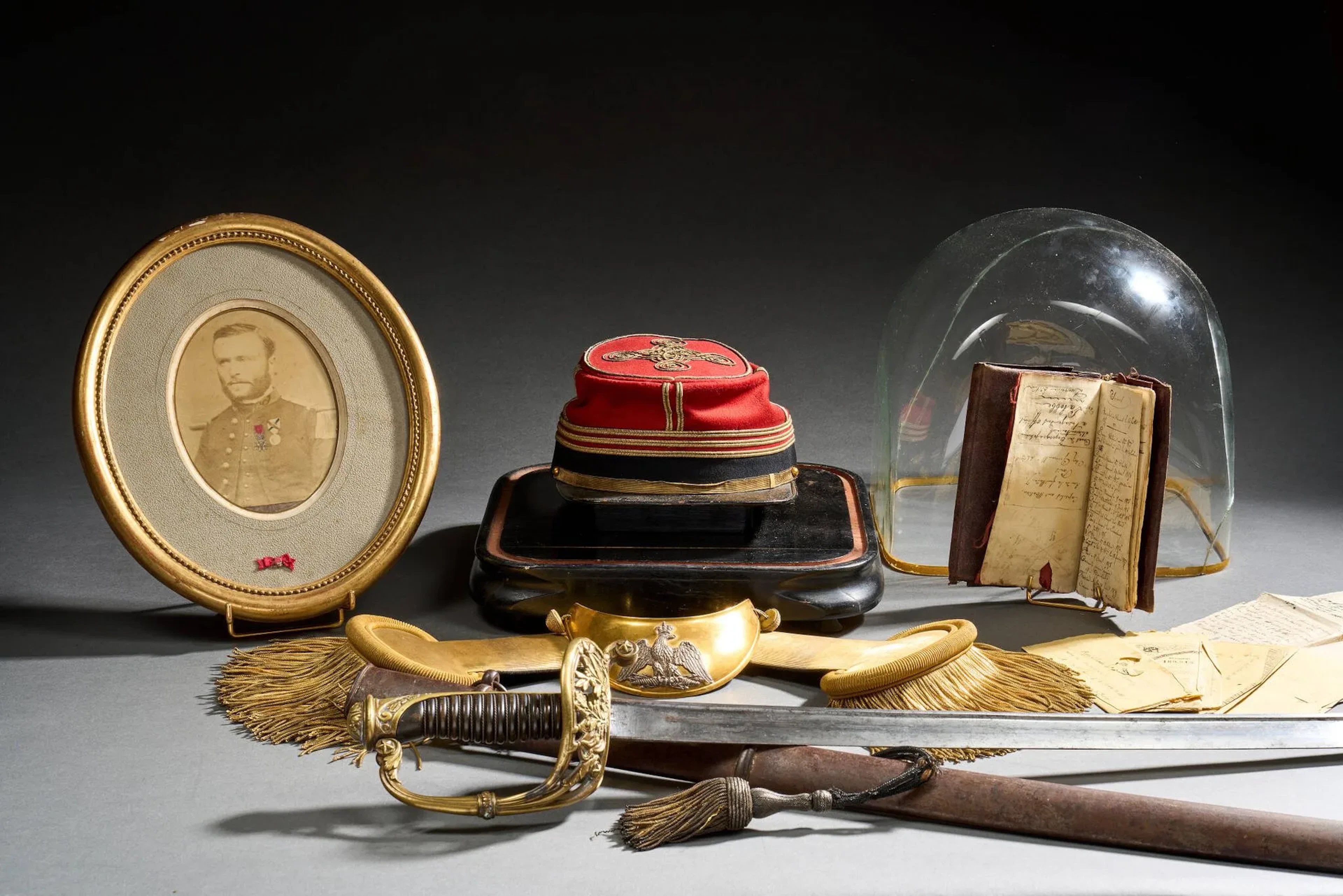 Ensemble ayant appartenu au Capitaine Charles-Marie-Adrieu Sorrel. Comprenant. -Kepi modèle 1860 au grade de Capitaine. Drap couleur garance et passementerie or. Visière type bec de canard estampée et bandeau intérieur en cuir fauve doré à la roulette. Intérieur doublé en soie et tampon de la maison Peisson Philippeville. Trou de mites sur le bandeau extérieur. -Paire d’épaulettes de la maison Herlouis, en passementerie or, doublé en drap de laine noir. Petites franges or et boutons au numéro 3 (probablement pour le troisième Zouave). -Portrait photographique avec médailles, rapportée pour la Légion d’honneur et rehaussée pour celle du Mexique. Passepartout avec ruban de la Légion d’honneur. États de services au dos (quelques erreurs aux dates). Hausse-col en laiton découpé à l’aigle argent. Complet de son drap de laine et boutons en passementerie. -Un portefeuille en maroquin impacté. Contient -Un feuillet manuscrit « Ce portefeuille trouvé quelque part après la bataille de froeschwiller par un paysan de Voerth, celui que portait au moment où il fut tué le Capitaine Sorrel ; il porte les traces d’une balle ; il a été remis à M. Sorrel père par le sous-préfet de Wisselbourg en mai 1872. » -six « traite du caissier payeur central du trésor public » de 500, 100, 500, 500, 1000 et 200 francs datés 1863 à 1868. Tous traversés par une balle. -Journal avec états de services et divers. -Sabre des chasseurs de Vincennes. Monture en bronze doré. Calotte ovale à longue queue décorée aux traits et couronne de lauriers. Arc de jointure percé pour le passage de la dragonne, dont se détachent deux branches formant la coquille à décors d’une couronne de lauriers sur fond de quatre fanions et étoiles. Quillon à enroulement à décor d’étoile. Fusée en corne filigranée. Lame droite à jonc par « Couleaux frères » à Klingenthal. Fourreau en fer à deux bracelets oxydations. On joint une dragonne à petites franges or (accidents). -Croix de guerre1914-1916 avec barrette de rappel et citation. -Globe en verre (accident).