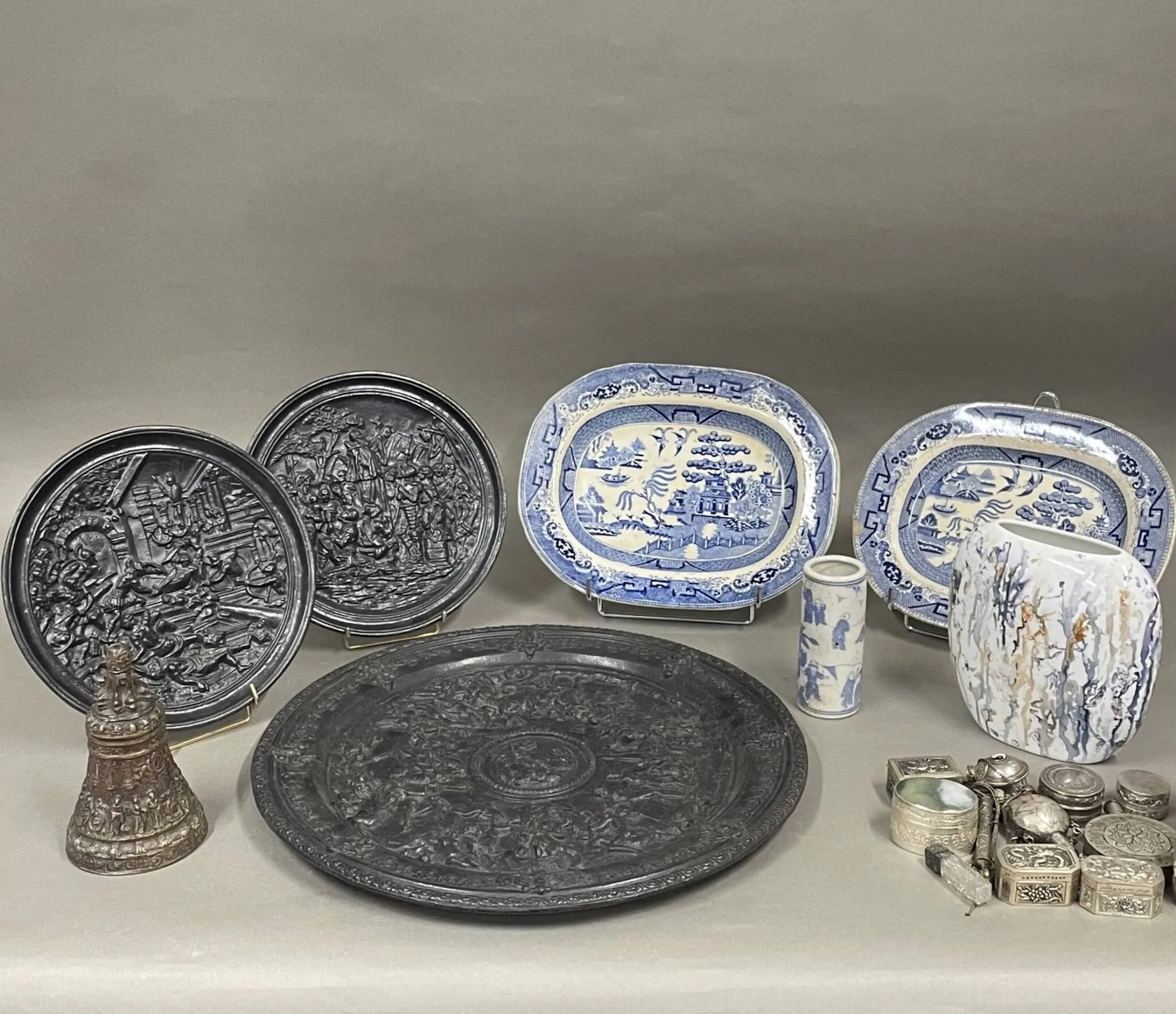 Lot de souvenirs comprenant -Un vase de section ovoïde en porcelaine, au décor de coulures colorés, monogrammé et daté "décor main / JB 11.2000" au dessous; H : 18,5 cm -Trois plats décoratifs en fonte patinée noire, à décors de scène de bataille antique, de messe de conquistadors et de banquet antique; le plus grand diamètre : 44,5 cm -Une cloche en bronze (manque le timbre) au décor tournant de personnages H : 16 cm -Deux plats de forme rectangulaire arrondie "Iron Stone China" en porcelaine au décor similaire de paysage animé en bleu, tamponnés au dessous (30 x 24,5 cm le plus petit), accidents -et un ensemble de petites boîtes, piluliers en métal argenté et divers dont vase cloisonné couvert, pot cylindrique en porcelaine de Chine (H : 13,5 cm) L'ensemble en l'état