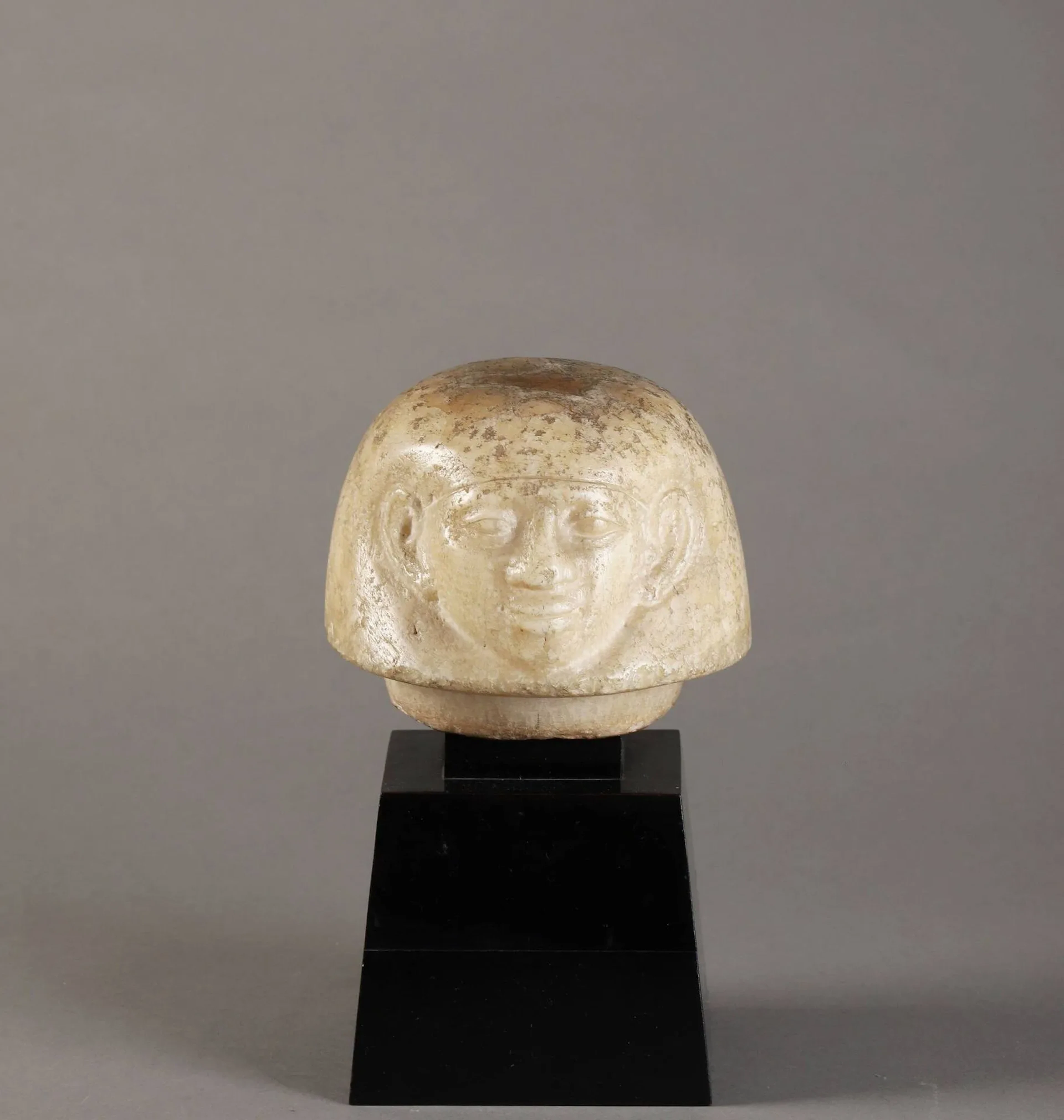 ÉGYPTE. Bouchon de vase canope en albâtre représentant le visage humain d'Amset, l'un des quatre fils d'Horus, coiffé d'une perruque dégageant les oreilles. IIe millénaire av. J.-C. Belle conservation malgré de petits éclats. H. 12 cm, D. 15 cm. Provenance : -Collection Kleimann, Paris. Probablement acquise dans les années 1950 et conservée dans la même famille depuis. Elle figure dans un inventaire de succession de 2002 - Vente Thomas enchères, Le Mans 18/02/2024 - Exceptionnelle collection de tableaux modernes et d'objets d'art Collection G K