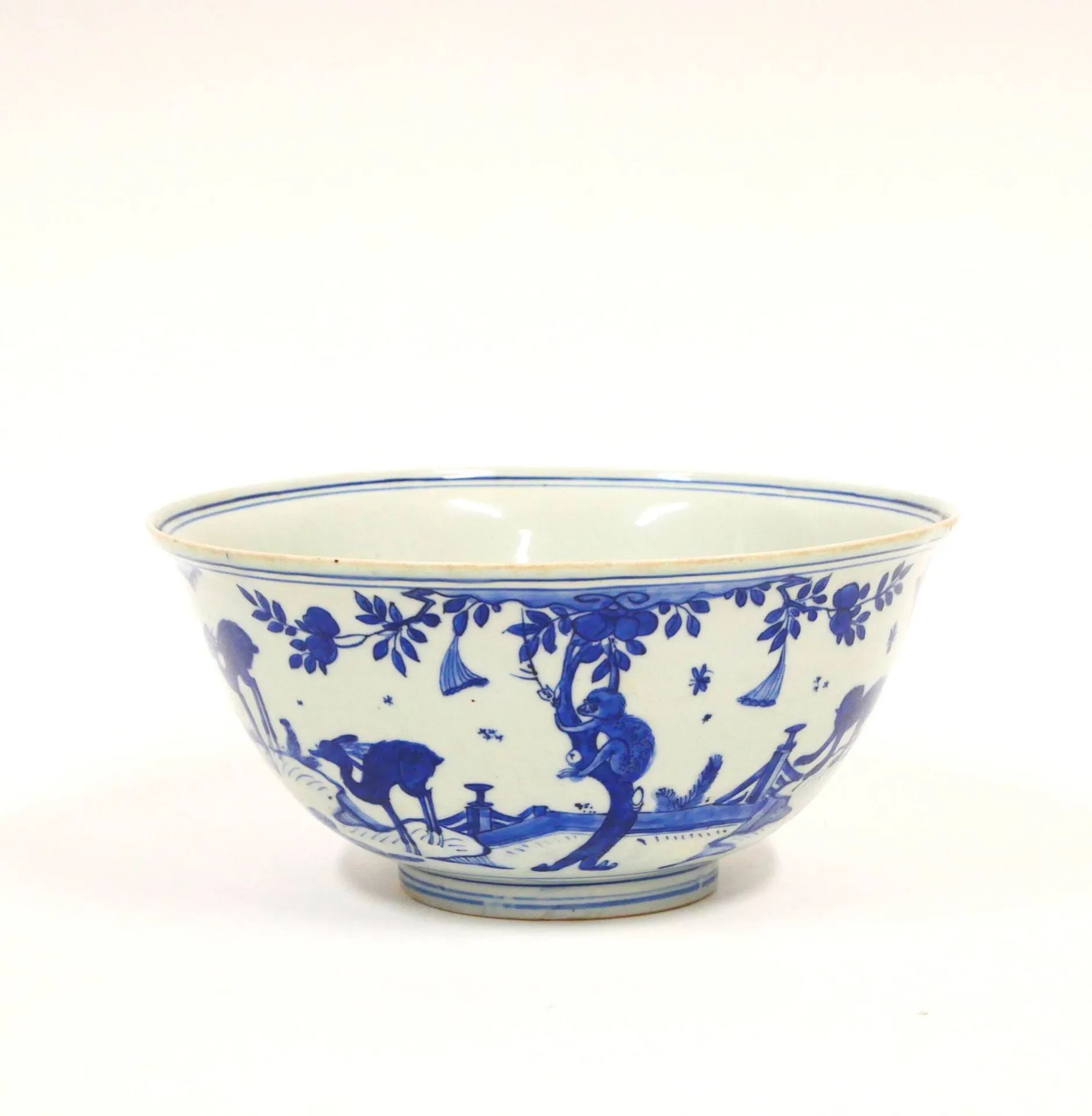 CHINE - Époque WANLI (1573-1620) Bol en porcelaine décorée en bleu sous couverte de singes, daims et abeilles sur une terrasse parmi les chrysanthèmes, un singe grimpant sur un pêcher pour cueillir une pêche. A l’intérieur, deux singes avec une abeille sous les pins. Au revers, la marque ‘Wanli nian zhi, yu tang jia qi’ (Objets du hall de jade, faut pendant la période Wanli). Minyao. (Fêlures de cuisson au talon). H. 11,5 cm Diam. 24,8 cm Le sujet du « singe et abeilles » est courant dans l’art chinois, symbolisant le vœu par homophonie: « que vous puissiez devenir marquis rapidement (mashang fenghou) ».