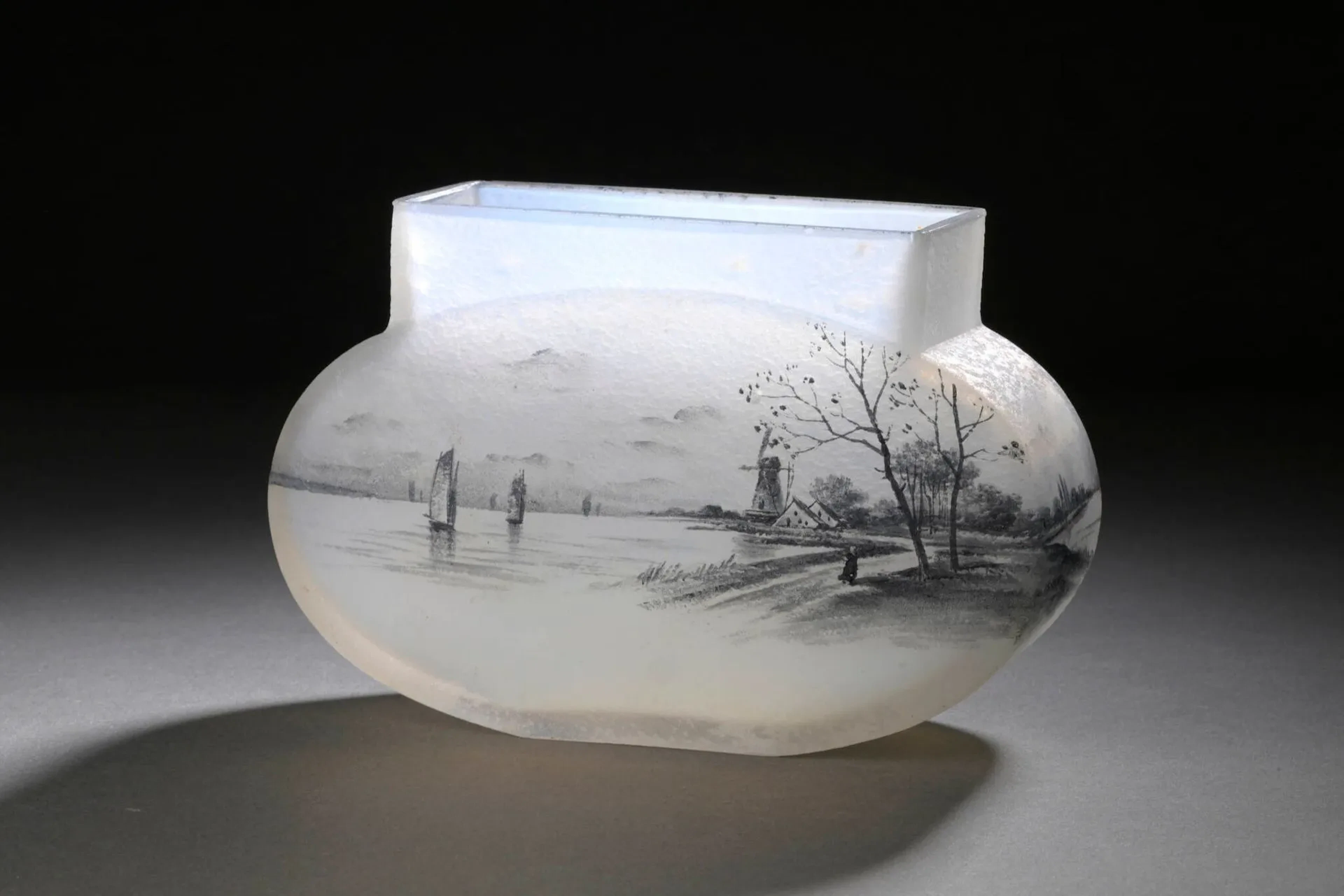 DAUM – NANCY Jardinière à corps ovoïde allongé épaulé et col quadrangulaire. Épreuve en verre blanc opalescent. Décor de voiliers et paysage lacustre sur fond d’un moulin. Émail en grisaille (infimes égrenures). Signée au revers. Haut. 11 cm – Long. 18 cm – Prof. 7 cm