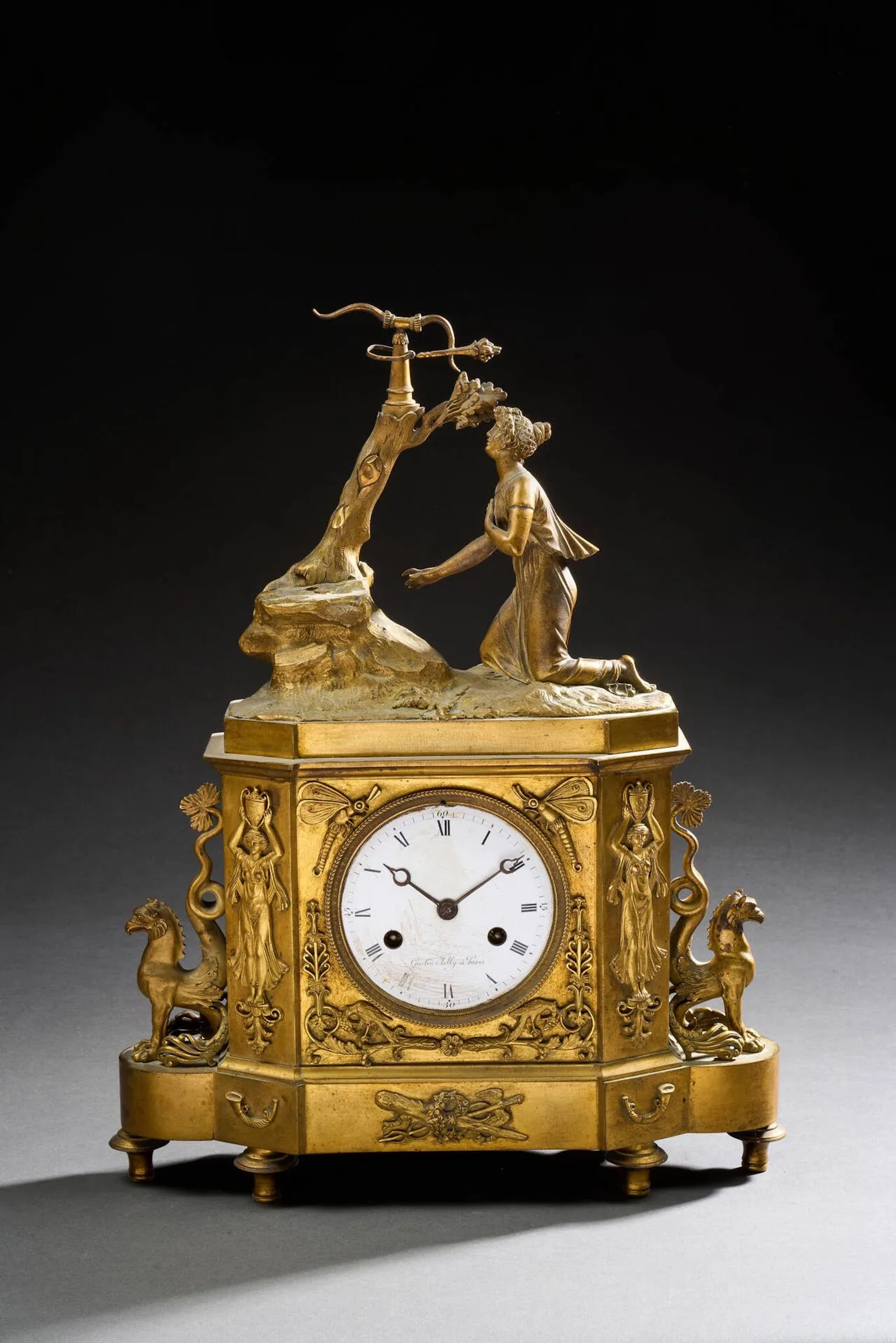 Pendule en bronze doré au sujet romantique figurant une jeune femme implorant à genoux devant un personnage (manquant) au pied d'un arbre. La base ornée d'animaux, de porteuses d'eau et flanquée de chimères. Cadran circulaire émaillé blanc en chiffres romains pour les heures et chemin de fer pour les minutes. Signé Gaston Jolly Paris. Platine arrière numérotée 305, suspensions à lamelles (manque le balancier). L'ensemble reposant sur six pieds. Manques. H : 42 cm.