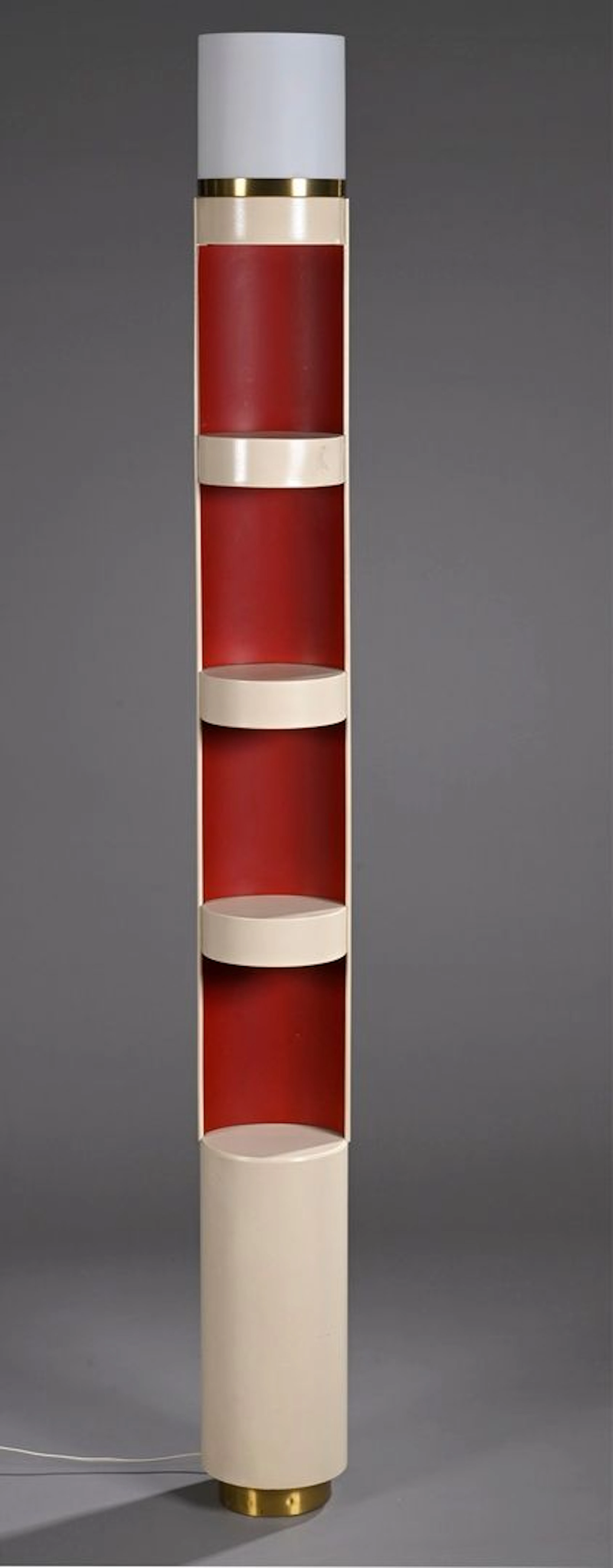 ATELIERS JEAN PERZEL Colonne lumineuse, modèle n° 966 du référencier de l’artiste, en laiton brossé et métal laqué crème et rouge à corps cylindrique et quatre tablettes formant niches intégrant un disque lumineux dans le fût et une lumière supérieure. Abat-jour tubulaire conique en verre blanc dépoli (petit enfoncement). Haut. 170 cm BIBLIOGRAPHIE : - Catalogue commercial, Paris, 1972, modèle référencé sous le n°966 et reproduit p. 13. - Jean-Jacques Wattel, Ateliers Jean Perzel, l’efficacité de la lumière, Éditions Louvre Victoire, Paris, 2023, pp. 211 et 216 pour des photographies datées de 1970 et 1974 montrant un modèle similaire présenté sur un stand de salon