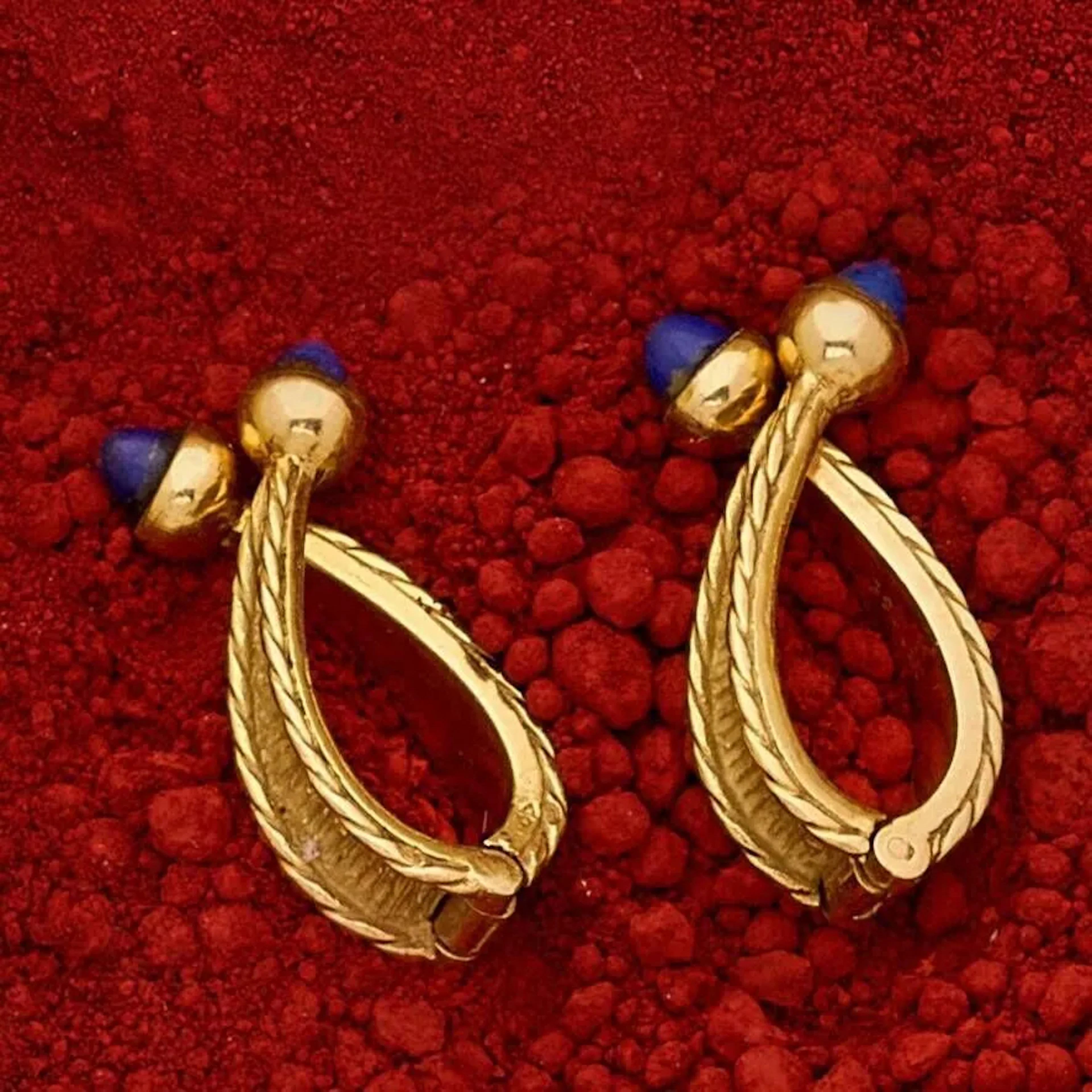 Paire de boutons de manchette en or 18k (750e), stylisant une boucle à décor texturé, bordés de torsades et terminés de cabochons de lapis-lazuli. (traces de colle). Travail français des années 1960/70. Poids brut : 11.30 g. Hauteur : 2.5 cm.