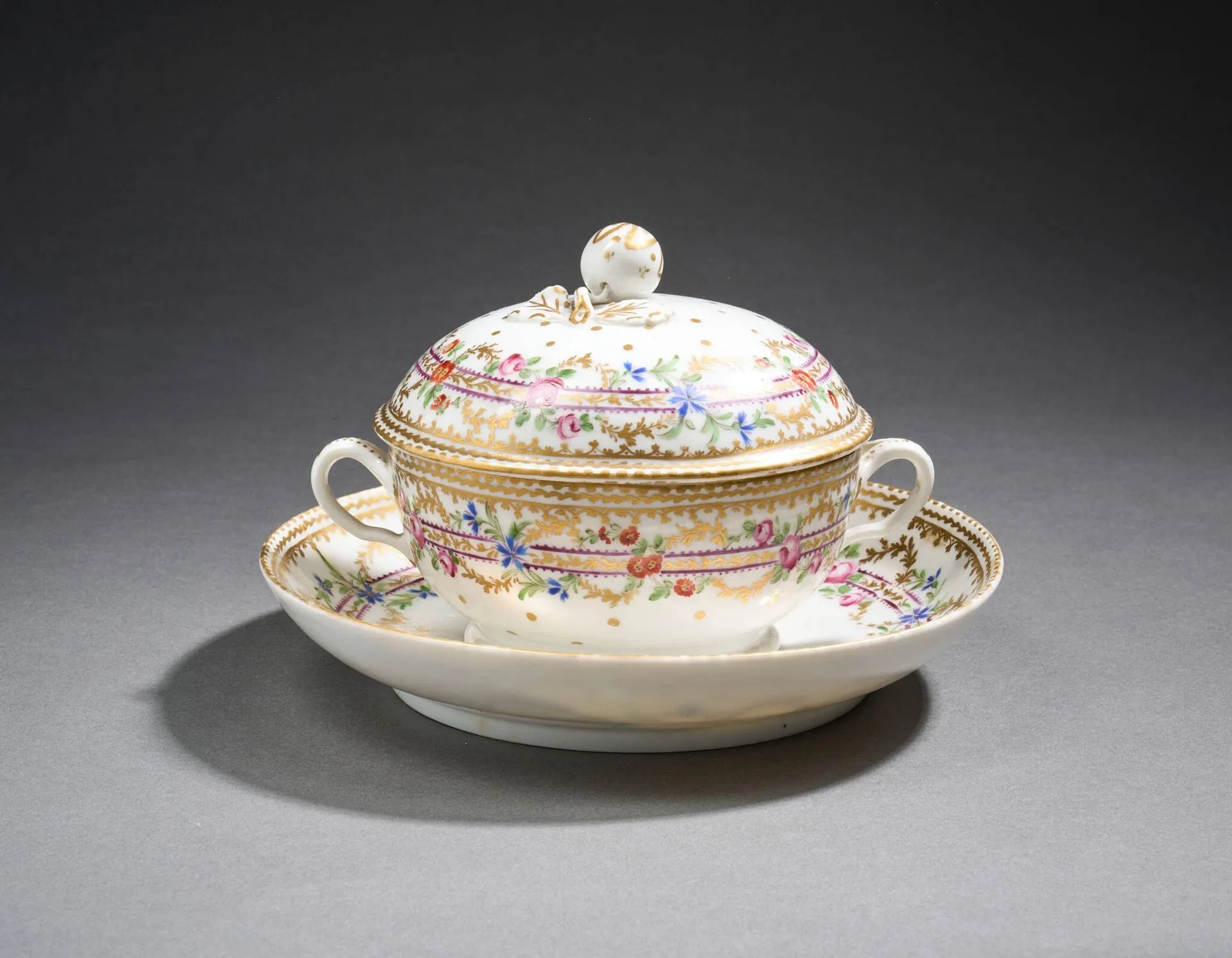 PARIS Bouillon couvert et son plateau en porcelaine à décor polychrome et or de frises de fleurs, dont barbeaux et roses, alternée de guirlandes végétales en or, dents de loup en bordure. Fêles et restaurations anciennes, usures de la dorure. Vincennes, manufacture Seguin, circa 1777-1788. Marque au lambel en bleu sous couverte au revers de la soucoupe. H. 13,5 x D. 19,5 cm.