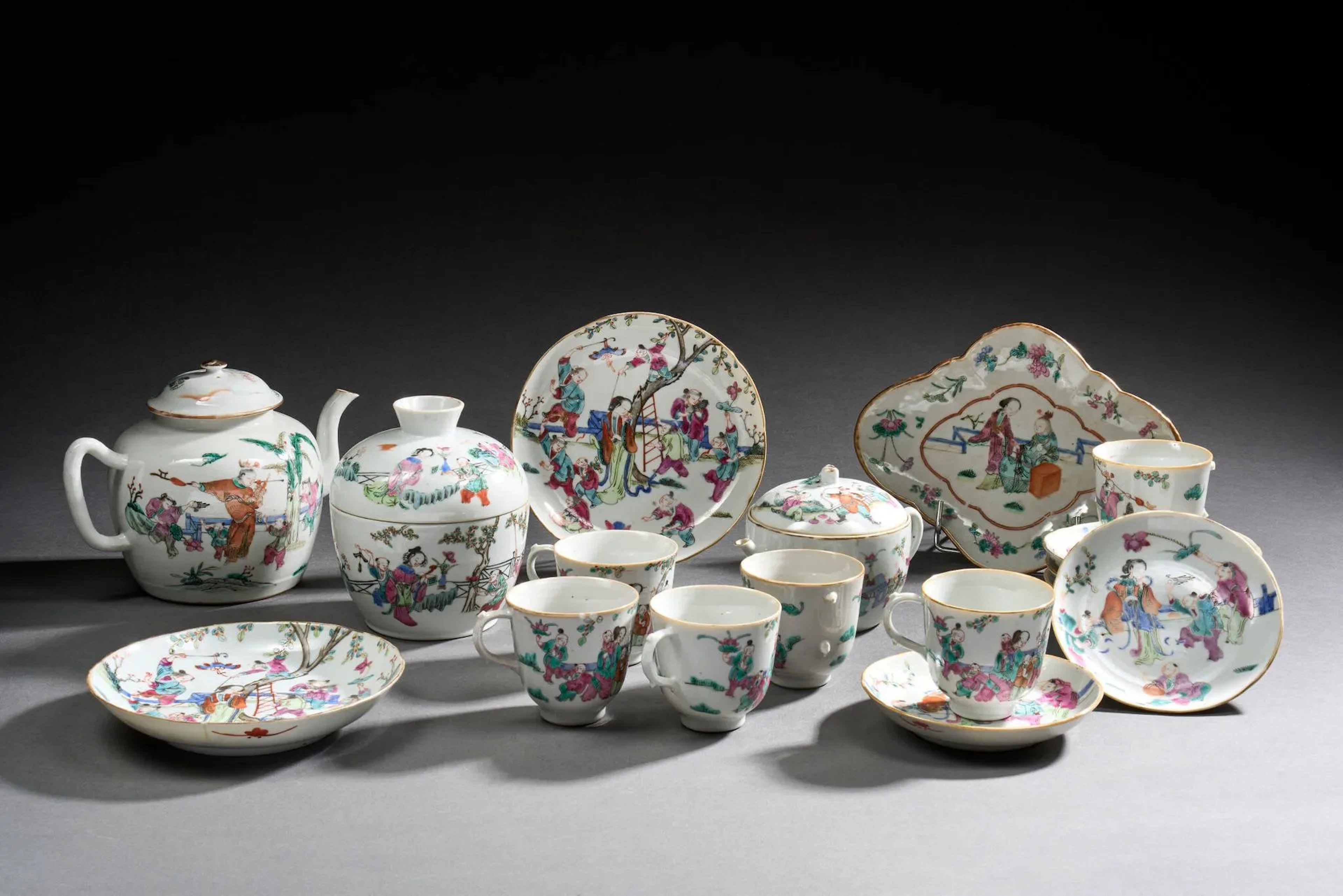 CHINE - Epoque TONGZHI (1862 - 1874) Ensemble en porcelaine émaillée polychrome dans le style de la famille rose comprenant une verseuse, une coupe polylobée, six tasses, six soucoupes, deux coupes, un pot couvert et une petite verseuse à décor d'immortels, femmes et enfants dans des jardins, certains enfants jouant de l'orgue à bouche et tenant des pierres sonores et des tiges de lotus. Au revers, la marque de Tonzghi. (Anses manquantes, égrenures et fêlures, couvercle rapporté) Diam. coupes 13,5 à 17,1 cm ; L. coupe polylobée 22,6 cm ; Diam. tasses sans anses 7,5 cm ; H. verseuses 10,4 à 15,7 cm ; H. boîte 14,8 cm