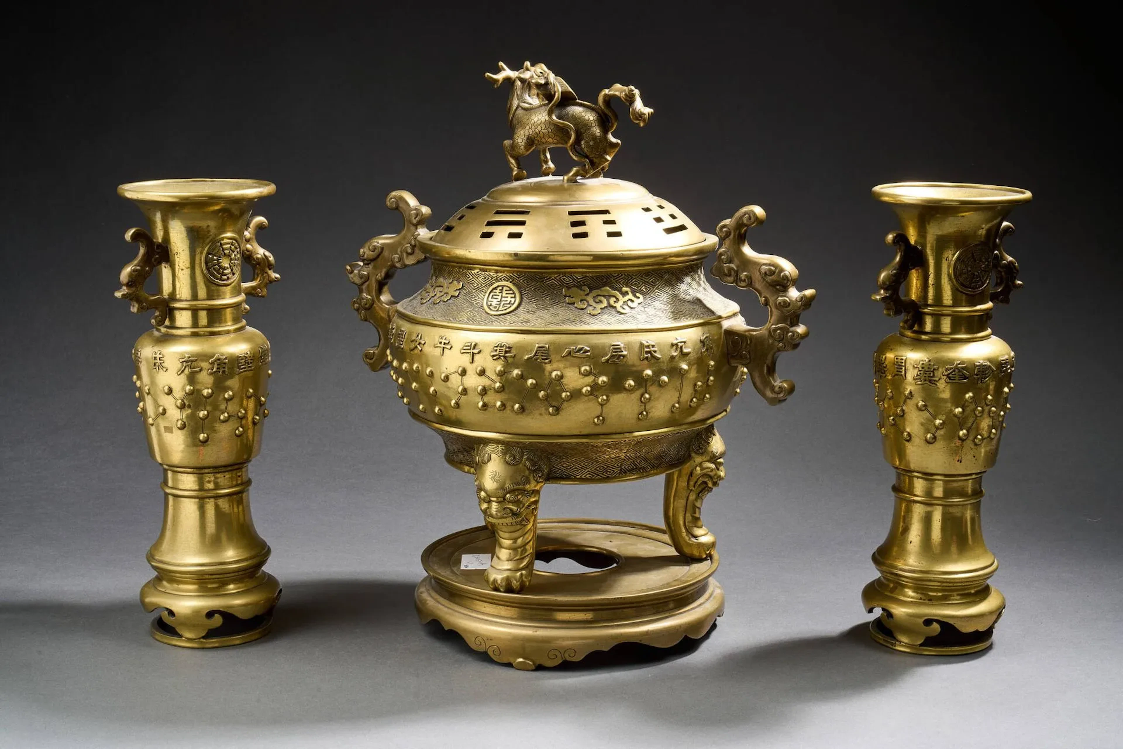 VIETNAM - Vers 1900 Garniture composée d'une paire de vases et d'un brûle-parfum tripode en bronze dépatiné, à décor sur la panse de constellations en relief et leur nom (28 loges nocturnes stellaires) Le brûle-parfum orné sur l'épaulement de nuages entourant un caractère Qian stylisé sur fond de motifs de leiwen, les pieds en forme de têtes de lions, les anses en forme de chilong formant nuages, le couvercle ajouré de trigrammes (bagua), la prise du couvercle en forme de qilin. La paire de vases comportant deux anses en forme de chilong formant nuages, le col orné de trigrammes (bagua). H. brûle-parfum 35 cm. H. avec socle 40 cm. H. vases 32cm