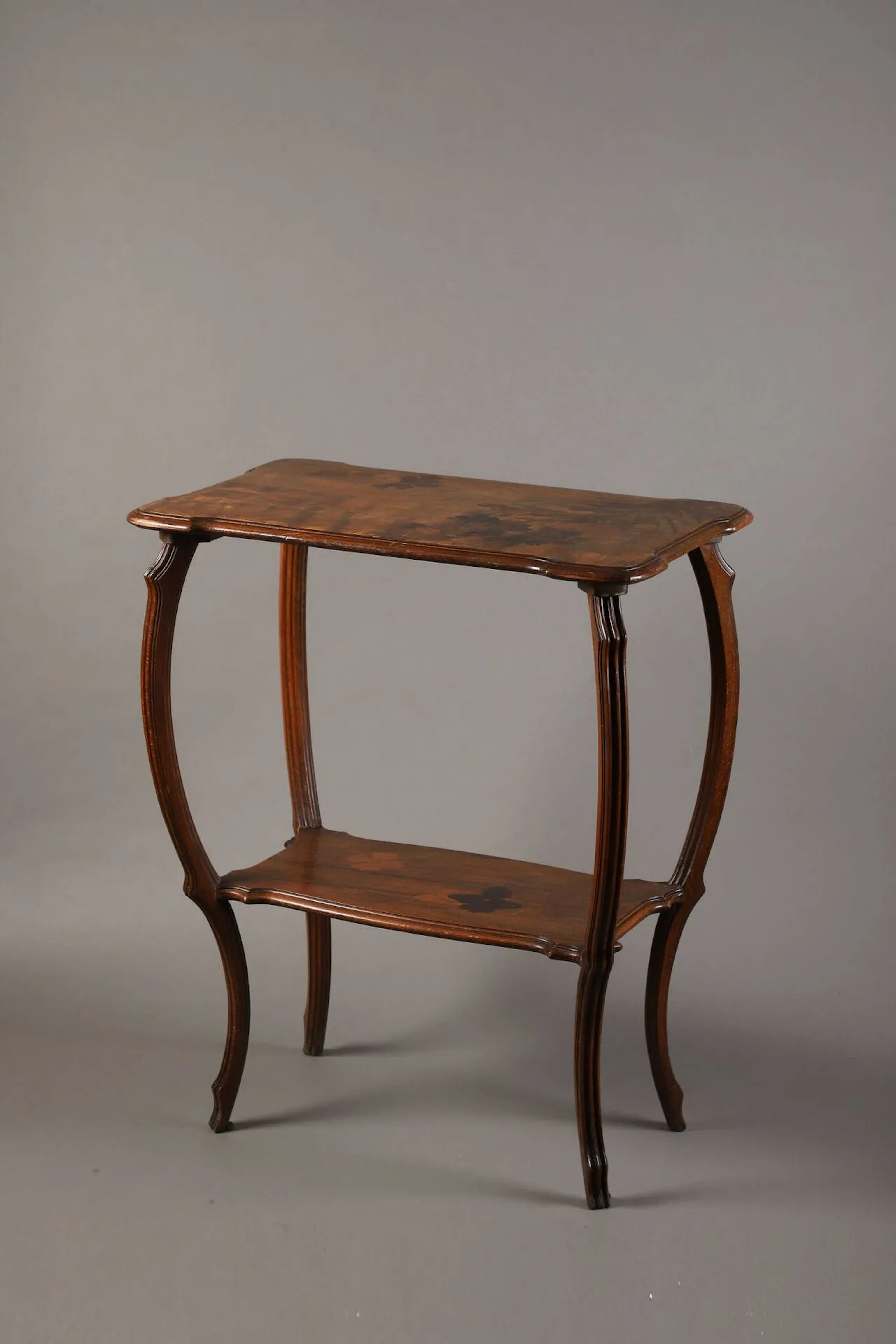 Émile GALLÉ (1846 – 1904) Table à thé en noyer mouluré et marqueterie de bois d’essences différentes à deux plateaux rectangulaires aux angles chantournés à décor de glycines pour le plateau supérieur et décor de deux fleurs pour le plateau inférieur. Piètement d’angle en léger retrait à jambes galbées rainurées (petits enfoncements, éraflures et taches). Signée dans la marqueterie du plateau supérieur. Haut. 74 cm – Long. 61 cm – Prof. 38 cm Bibliographie : Alastair Duncan & Georges de Bartha, Gallé Furniture, Antique Collectors’ Club, Woodbridge, 2012, table de forme similaire à notre modèle référencée sous le n°69 et reproduite p. 154.