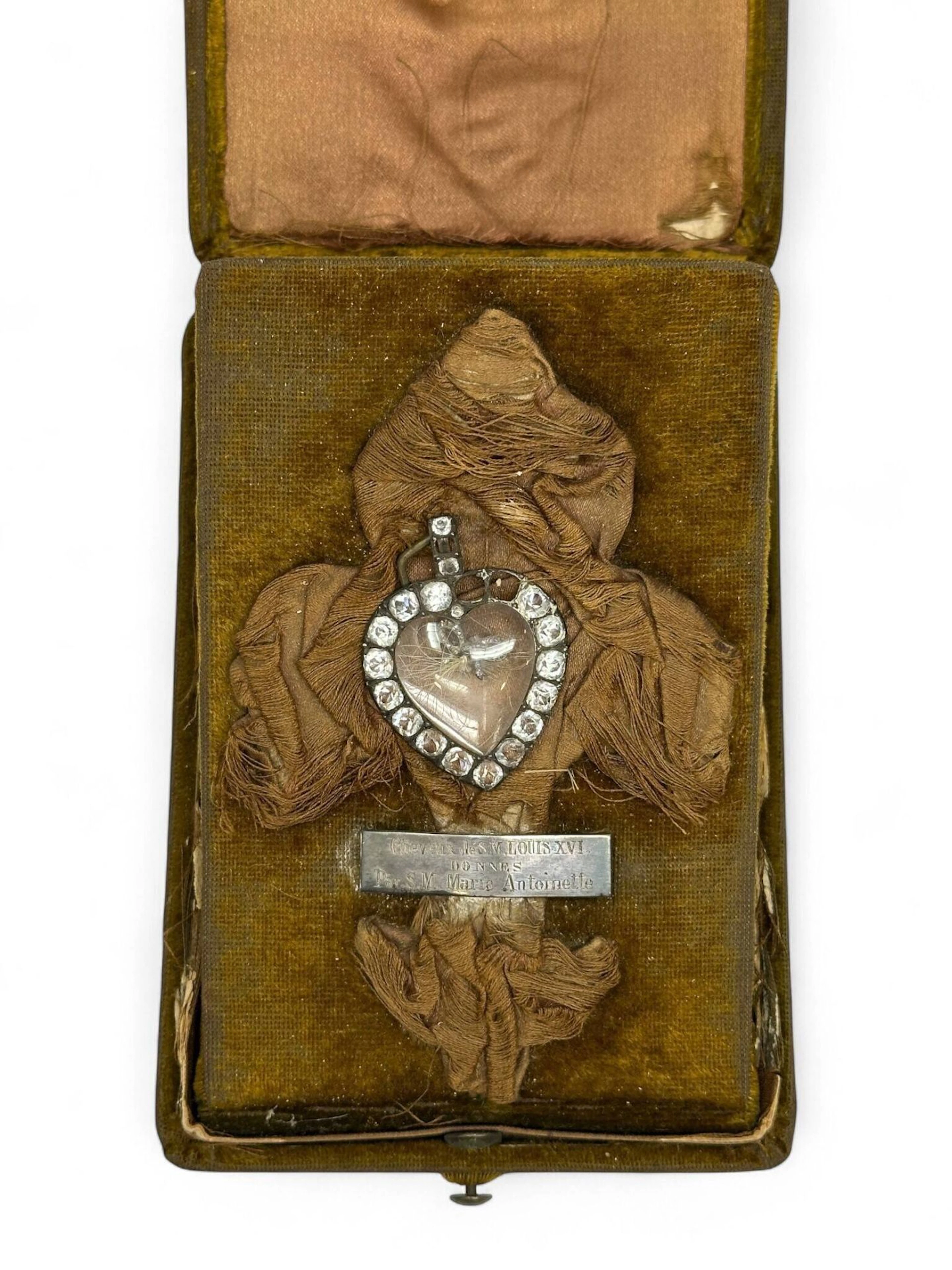 PRÉCIEUSE RELIQUE DU ROI LOUIS XVI Médaillon pendentif en forme de cœur en argent (800 millièmes) et or bas-titre serti de strass (trois manquants), la partie centrale en cristal bombé s’ouvrant à charnière renferme une mèche de cheveux blancs nouée et retenue par un nœud rubané en argent serti de pierres blanches. Le médaillon est conservé dans un écrin rectangulaire tendu de velours vert et est présenté au centre d’une fleur de lys en soie brune, accompagné d’une plaque en argent gravée « Cheveux de S.M. Louis XVI / donnés / par Marie Antoinette ». H 4,3 x L. 3,2 cm. Poids brut : 9,3 g (médaillon). Provenance Château de La Noë-Sèche (Côte d’Armor), ancienne collection du vicomte Jules-Marie Armand Cavelier de Cuverville (1834-1912), vice-amiral, chef d’Etat-Major de la Marine, Sénateur du Finistère entre 1901 et 1912. Exposition Marie-Antoinette et son temps, Galerie Sedelmeyer, 4 bis, rue de la Rochefoucauld, Paris, 1894, sous le n° 44. Littérature Cette relique est mentionnée dans la Revue des Beaux-Arts, dans l’article consacré à l’exposition « Marie-Antoinette et son temps », p. 117 : « Voici ceux que nous avons notés un peu au hasard, car tous seraient à citer, le choix, parmi les objets offerts, ayant été fait avec la plus grande circonspection et le goût le plus parfait par les organisateurs de l’Exposition… Un rouet tourné par Louis XVI ; un cœur en diamant avec cheveux du roi ; un rouet fait par Louis XVI pour Marie-Antoinette et donné par elle à une parente du propriétaire - à M. Le vicomte de Cuverville ».