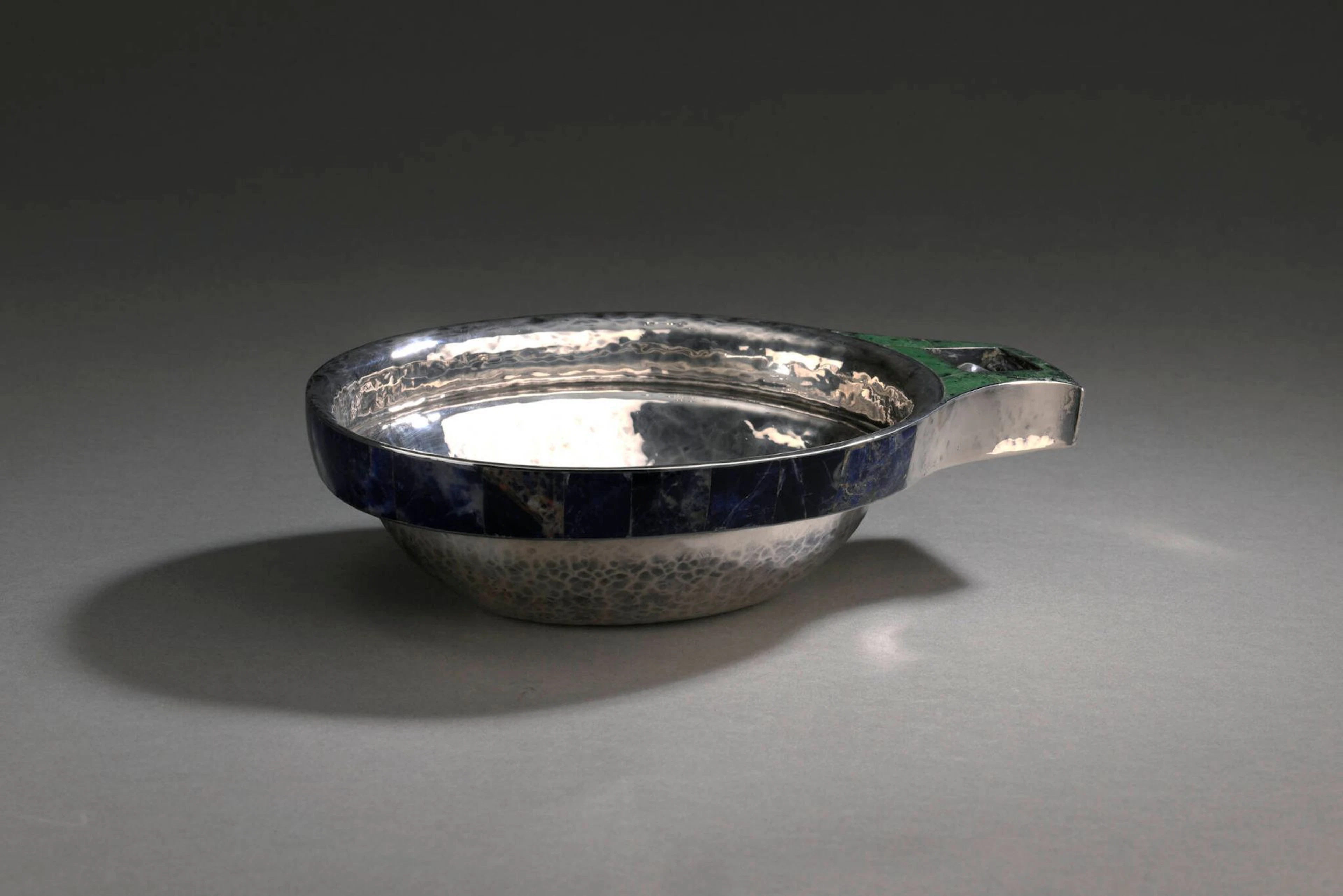 GOUDJI (né en 1941) [Guy Georges Amachoukeli dit] Coupe en argent (925) martelé, le col souligné d'incrustations de plaquettes de lapis lazuli et la prise à décor ajouré d'un cerf stylisé dans un encadrement de plaquettes de jade néphrite. Signée sur le dessous, marque de l'artiste et marqué PARIS Poinçon de titre: 925; Poinçon d'orfèvre Poids brut: 587,5 g H. 6,2 - L. 23 ; P. 15,6 cm Légères traces d’usage et légers éclats ponctuels.