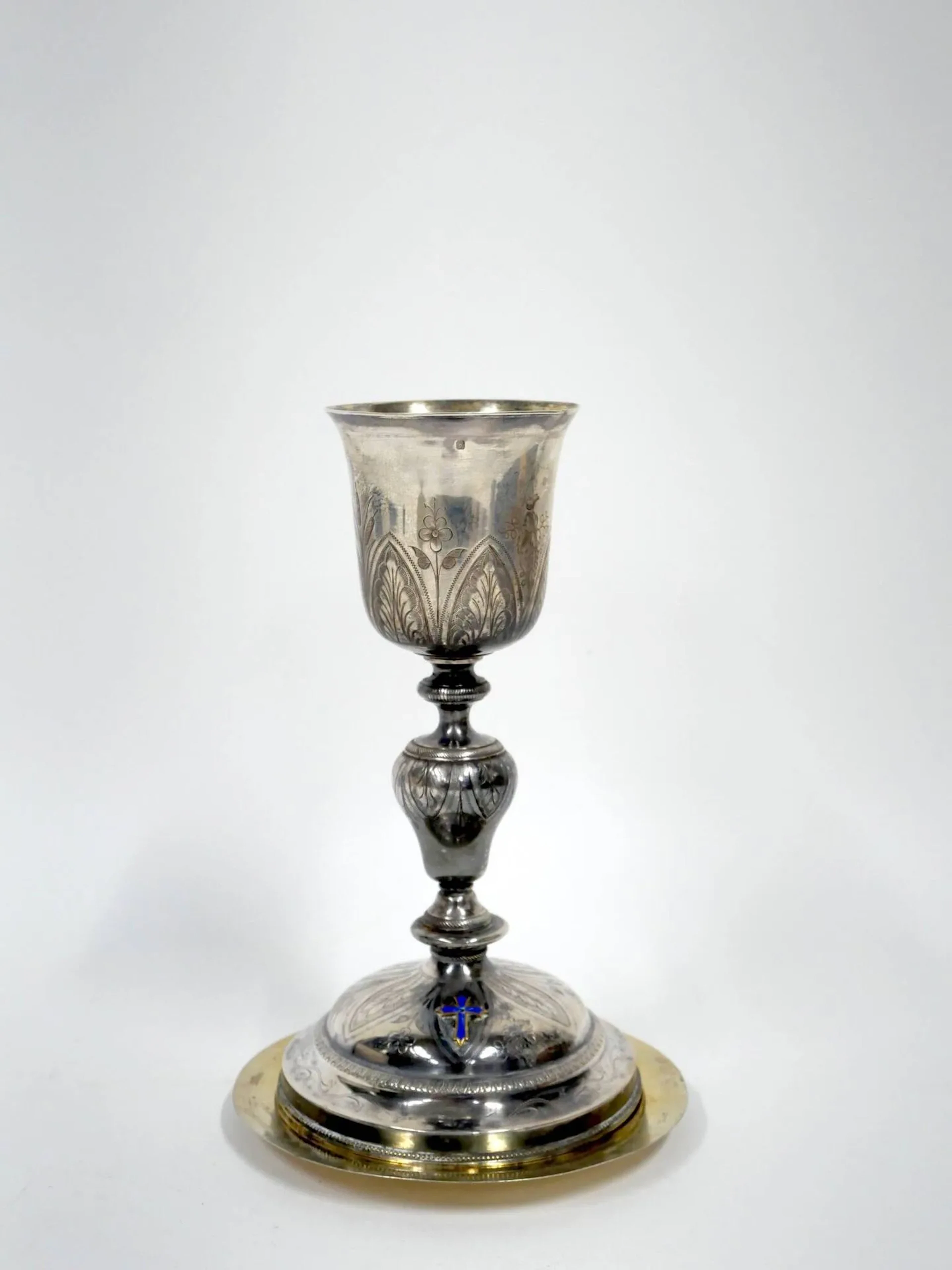 Calice et un patène en argent (950), métal et vermeil entièrement gravé de feuilles lancéolés, d'épis de blé, de fleurs et de roseaux, le fût à balustre et à bagues, le piédouche circulaire surmonté d'une croix émaillé bleue (accidents). Le patène (rapporté) ouvragé d'une croix sur fond amati et de l'inscription "IHS". Orfèvres : illisibles Poids brut de l'ensemble : 355 g ; Hauteur : 22 cm Manque à la dorure et accidents.