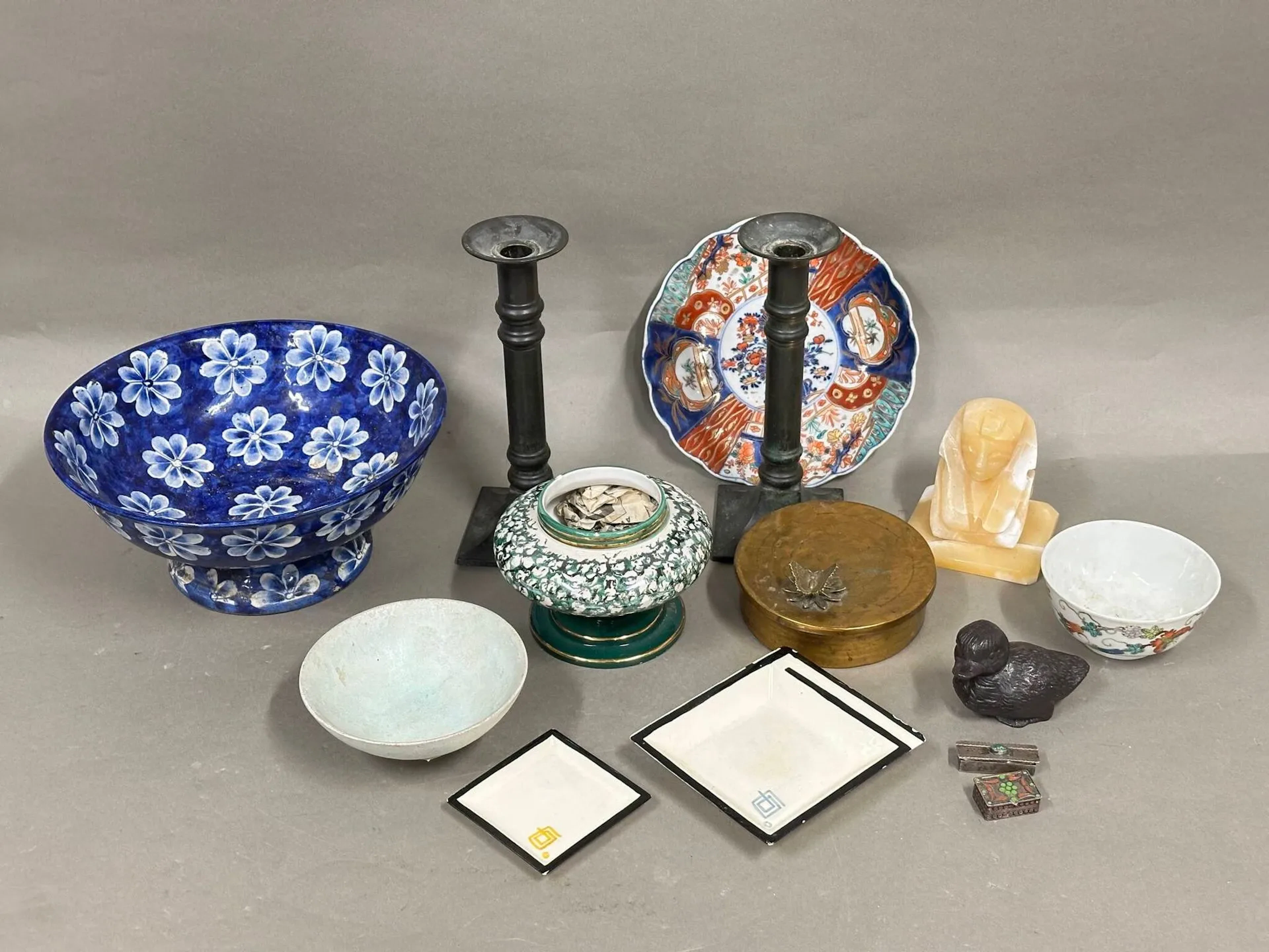 Lot comprenant : Une coupe en porcelaine émaillée bleue à décor de fleurs Une assiette Imari en porcelaine à décor floral Un coupe monogrammé LB Un bol en porcelaine de Chine à décor de rinceaux de fleurs et de papillons portant une marque au dos. Une coupe en porcelaine blanche et verte numérotée 5/297 et signée "BASSAMO" Une paire de flambeaux "CHRISTIAN DE BEAUMONT" Un canard en bronze patiné Deux cendriers signés "MARC HENRI" Un serre livre sphynx en pierre dure Une boite Mappin & Webb en laiton à décor de papillon Deux boites en métal et émail En l'état.
