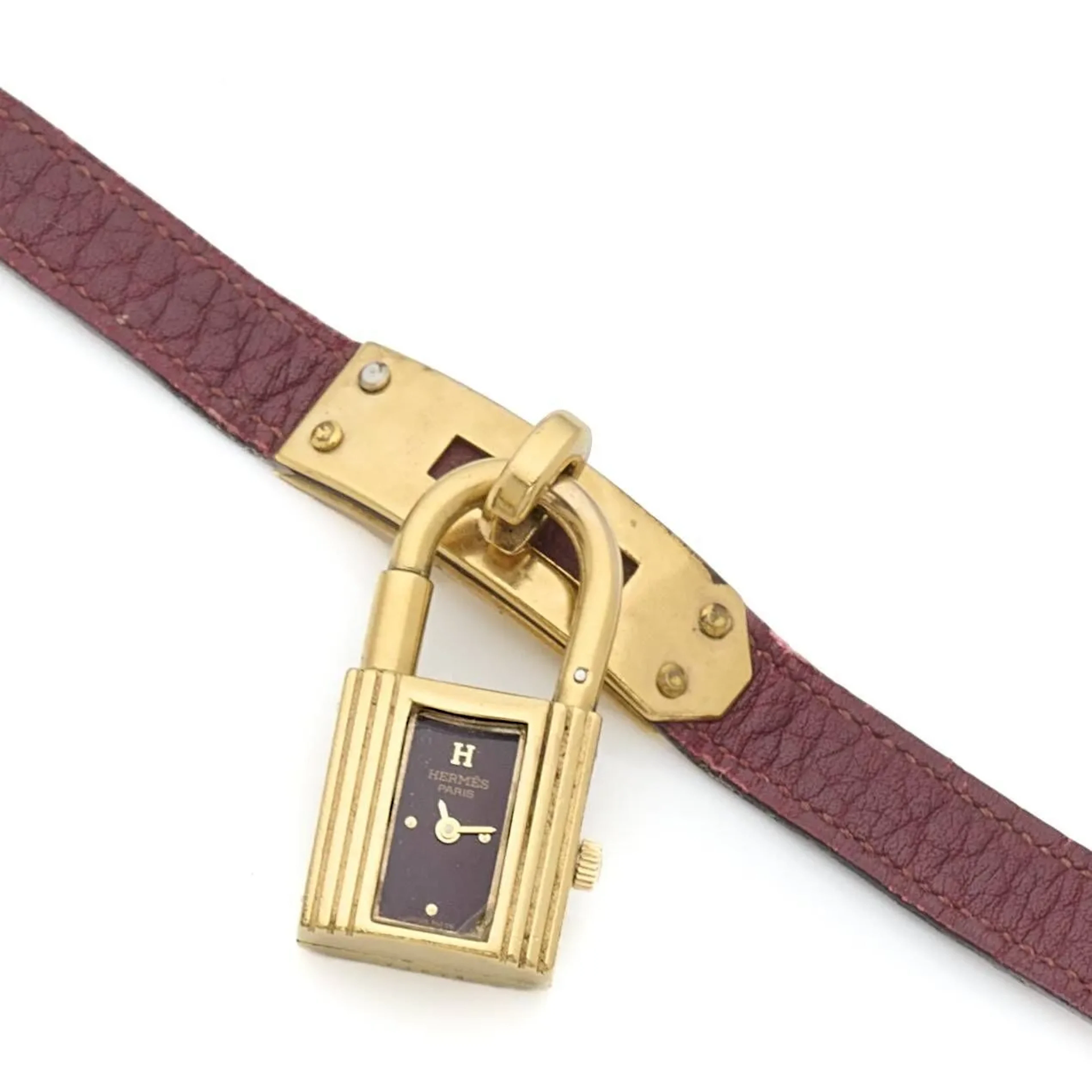 HERMÈS KELLY Montre bracelet en métal plaqué or 10 microns pendante façon cadenas, cadran bordeaux, index en applique dorés, aiguilles dorées. Boitier numéroté 73674. Mouvement à quartz signé Hermès base ETA. Bracelet en cuir marron, insert du cadenas en métal plaqué or, boucle ardillon en métal doré. Mouvement non fonctionnel lors de l’expertise, sans garantie de fonctionnement futur et d'état des pièces. Rayures et marques, accident au verre. Vers 1990