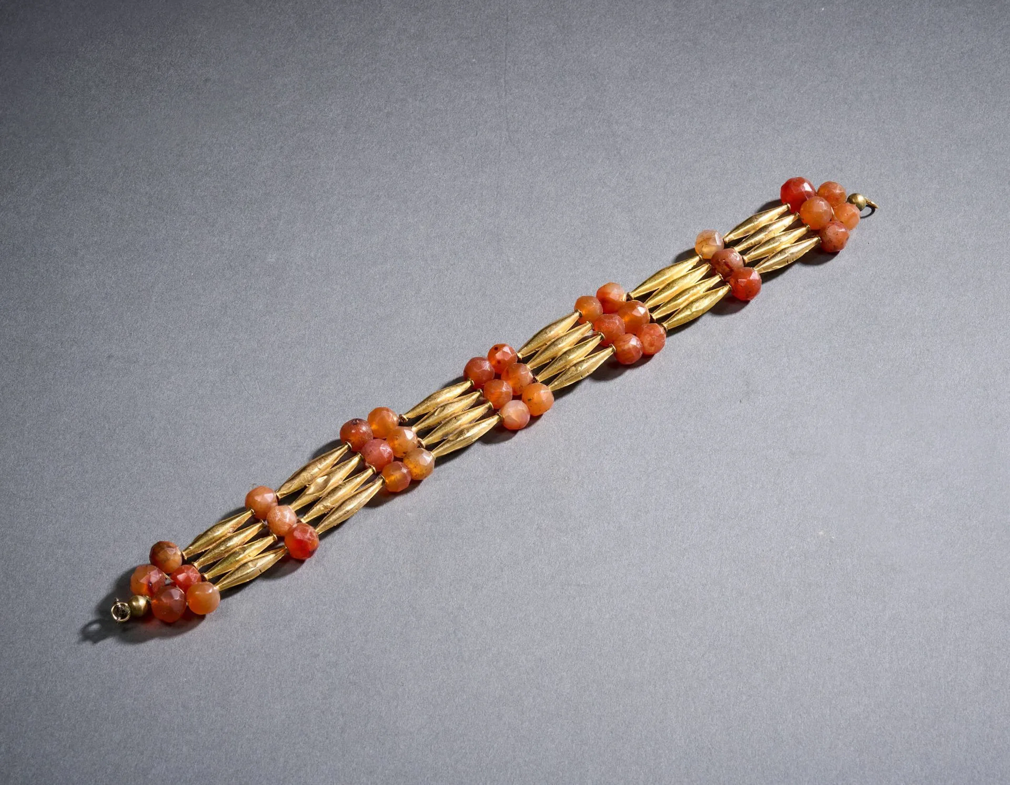 Collier draperie en métal doré et cornaline, à décor alterné de perles en cornaline facettées sur trois rangs et de perles tubulaires en or de forme légèrement oblongue sur quatre rangs, le fermoir en or bas titre rapporté. Petits chocs de l’or, le fermoir cassé. Travail égyptien du XIXe siècle, dans le goût antique. Sans poinçon apparent. L. 30 x H. 3. Poids brut : 60,0 g.