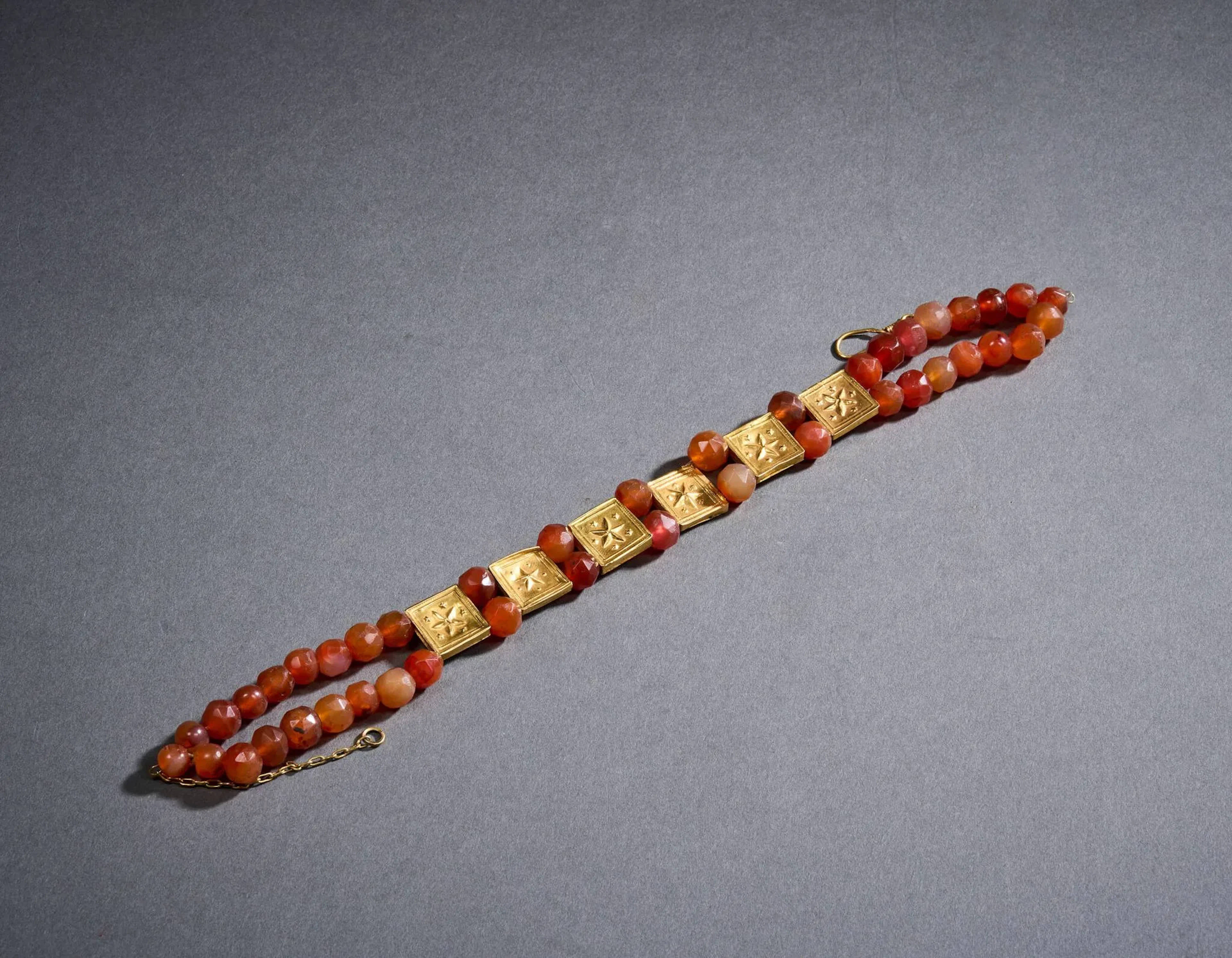 Collier draperie en or (585 millièmes) et cornaline, à décor alterné de perles en cornaline facettées sur trois rangs et de carrés en or à décor repoussé de motifs géométriques stylisés, se fermant par une chaînette et un fermoir en or (585 millièmes), rapportés. Petits chocs. Travail égyptien du XIXe siècle, dans le goût antique. Porte un poinçon de contrôle égyptien. L. 39 cm. Poids brut : 51,1 g.