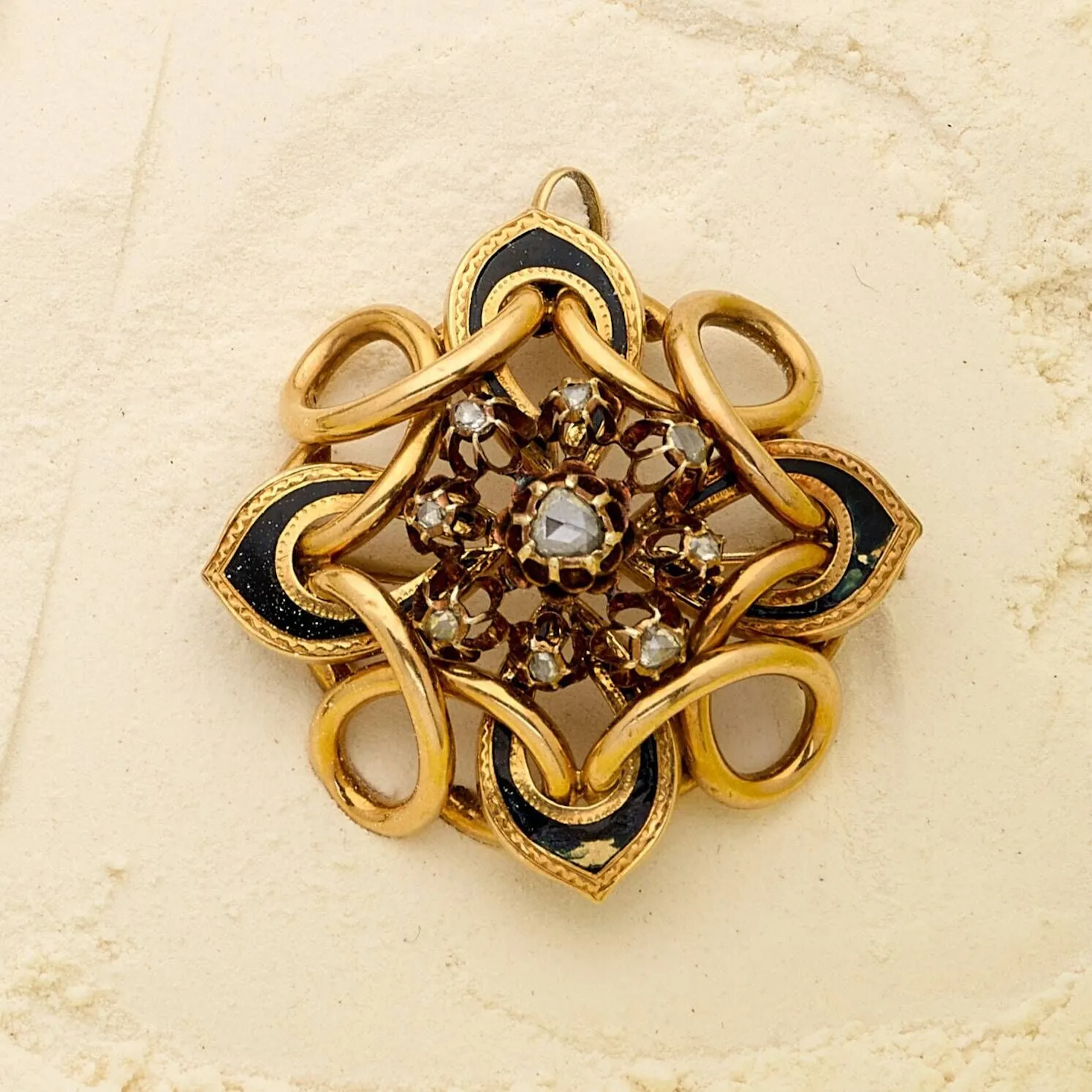 Broche pendentif en or 18k (750e), à décor d'entrelacs rehaussés d'émail noir, centrée d'un motif floral orné de diamants taillés en rose dont un plus important au centre. (manques à l'émail, enfoncements, remontage en pendentif). Travail français d'époque Napoléon III. Poids brut : 16.60 g. Diamètre : 4.5 cm.