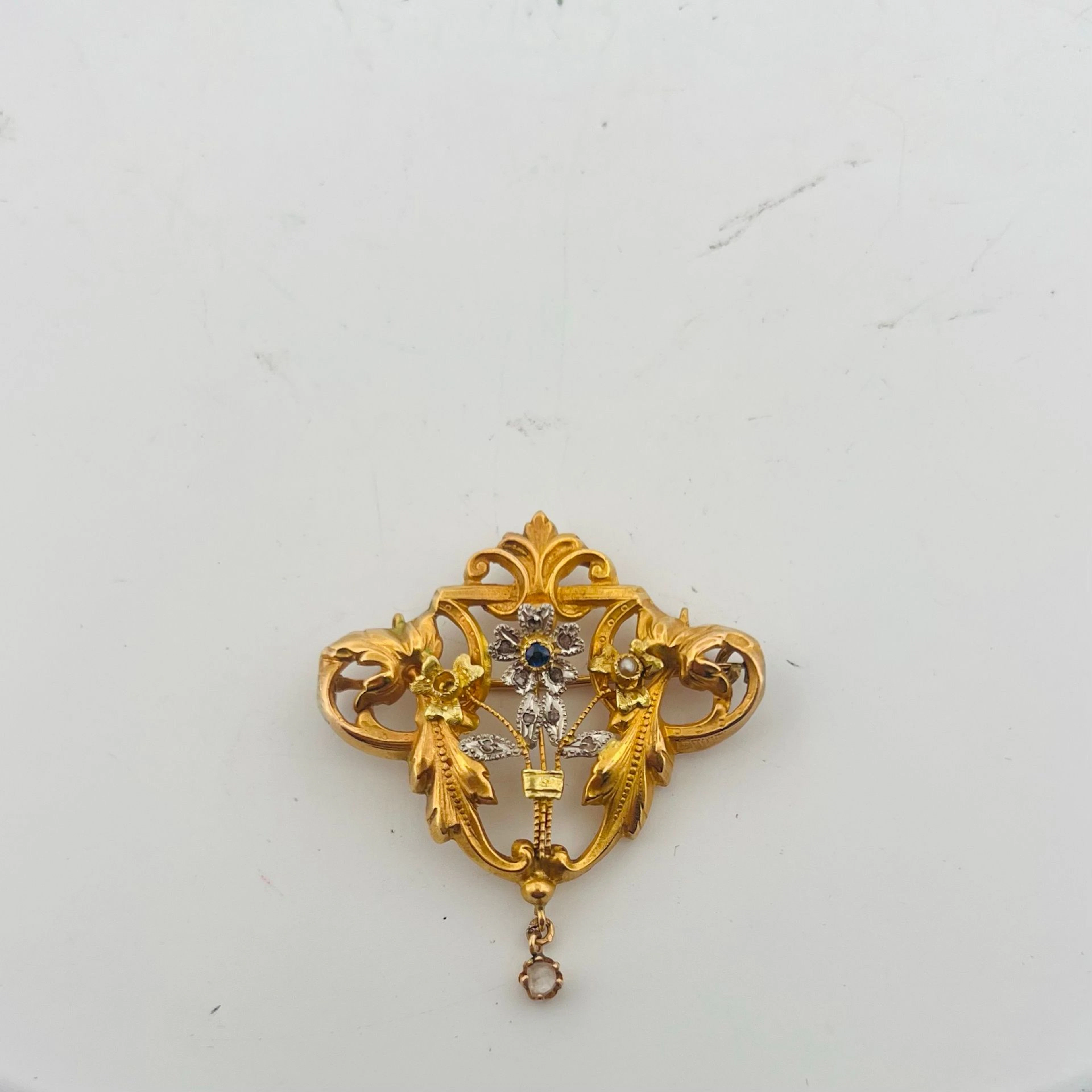 Broche en or 18K (750) à décorde volutes feuillagées, centrée d'un motif floral piqué 'une pierre bleue et se terminant par une pampille ornée d'une pierre blanche. Poids brut : 2.96 g. (manques, réparations, système pour être porté en pendentif)