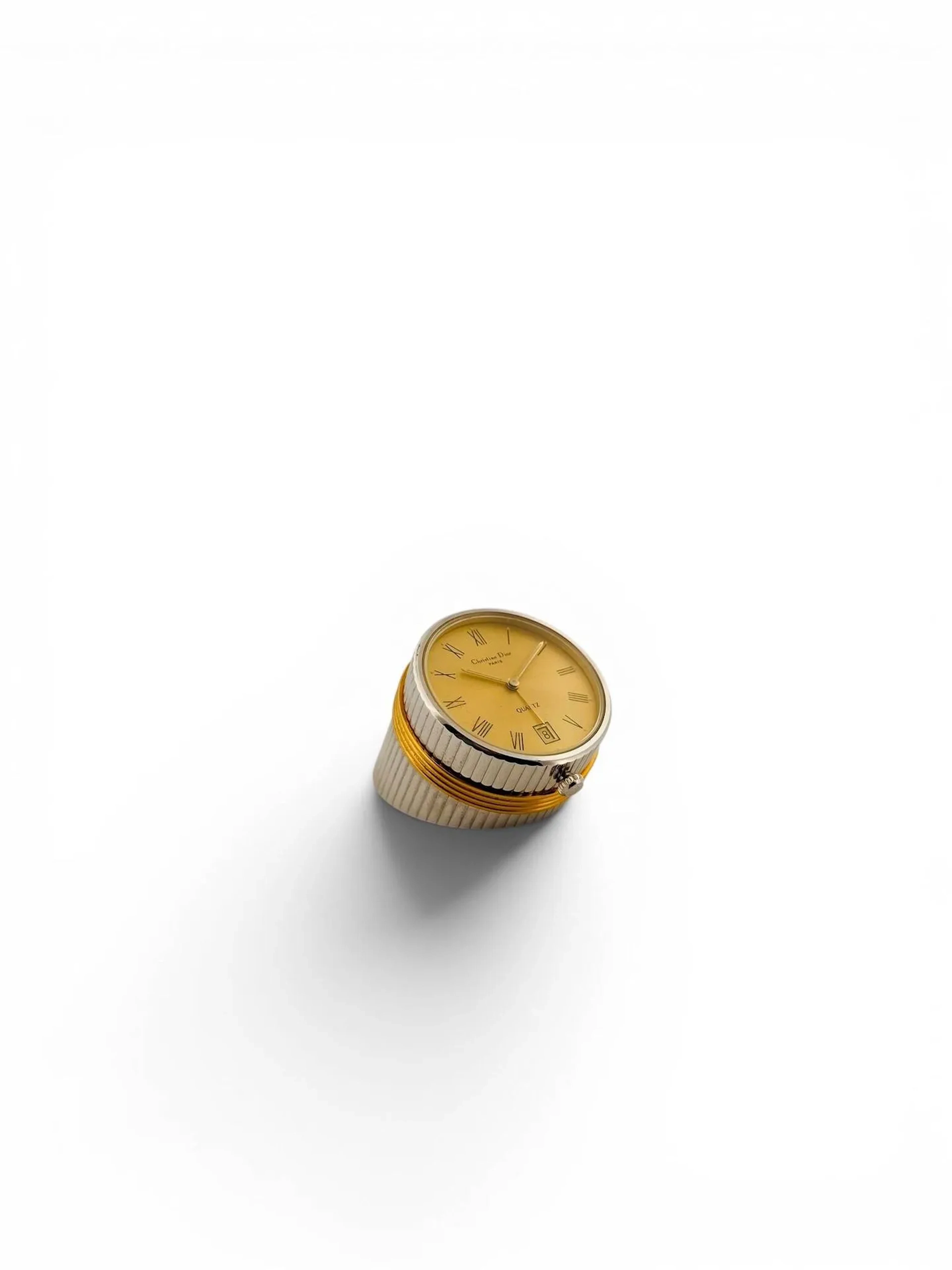 Christian DIOR Pendulette de table en métal argenté et doré à décor de cannelures annelées en partie haute, cadran champagne à chiffres romains noirs, guichet dateur à 6 heures. Fond de boîtier signé et chiffré "37-20-01 50229" Mouvement quartz H. 3 - Diam. 3,4 cm Dans l'écrin d'origine