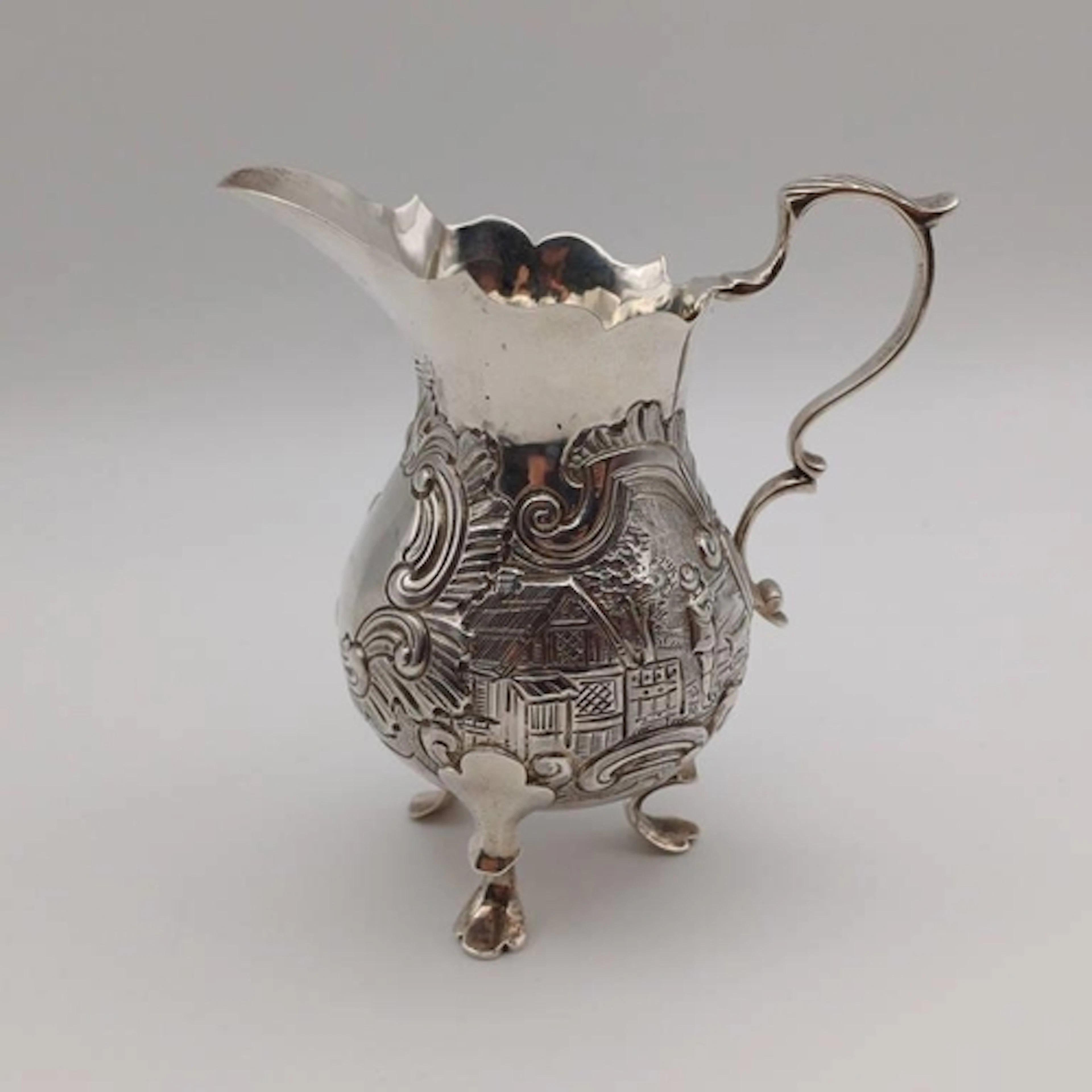 A VICTORIAN SILVER CREAM JUG WITH EMBOSSED VILLAGE SCENE DEC...